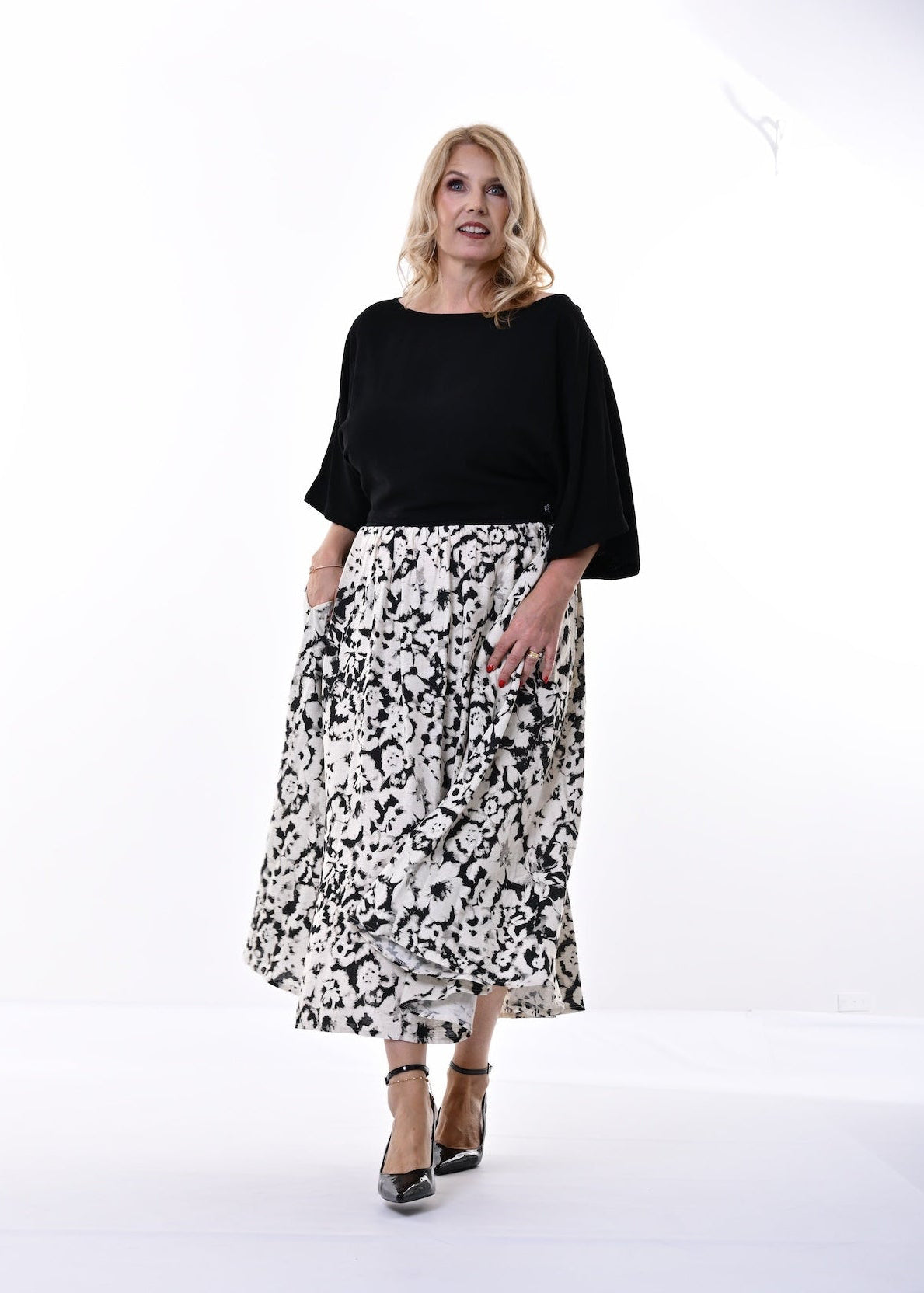 A woman modeling a black short-sleeve top and a black and white floral print midi skirt, paired with black high heels, against a plain white studio background.