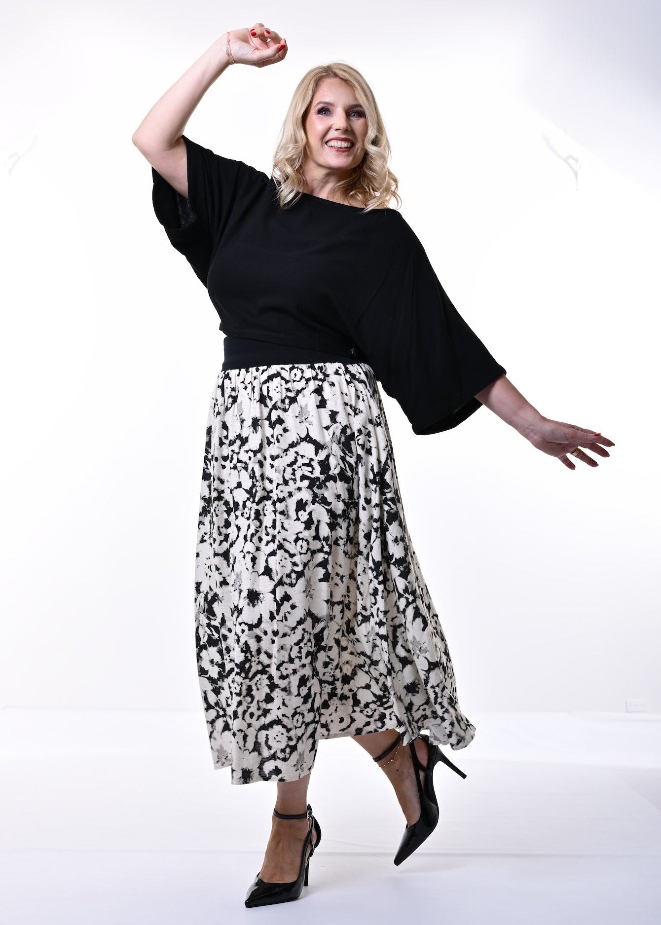 A woman modeling a black 3/4 length sleeve top and a black and white abstract patterned midi skirt, paired with black heels, against a plain white studio background.