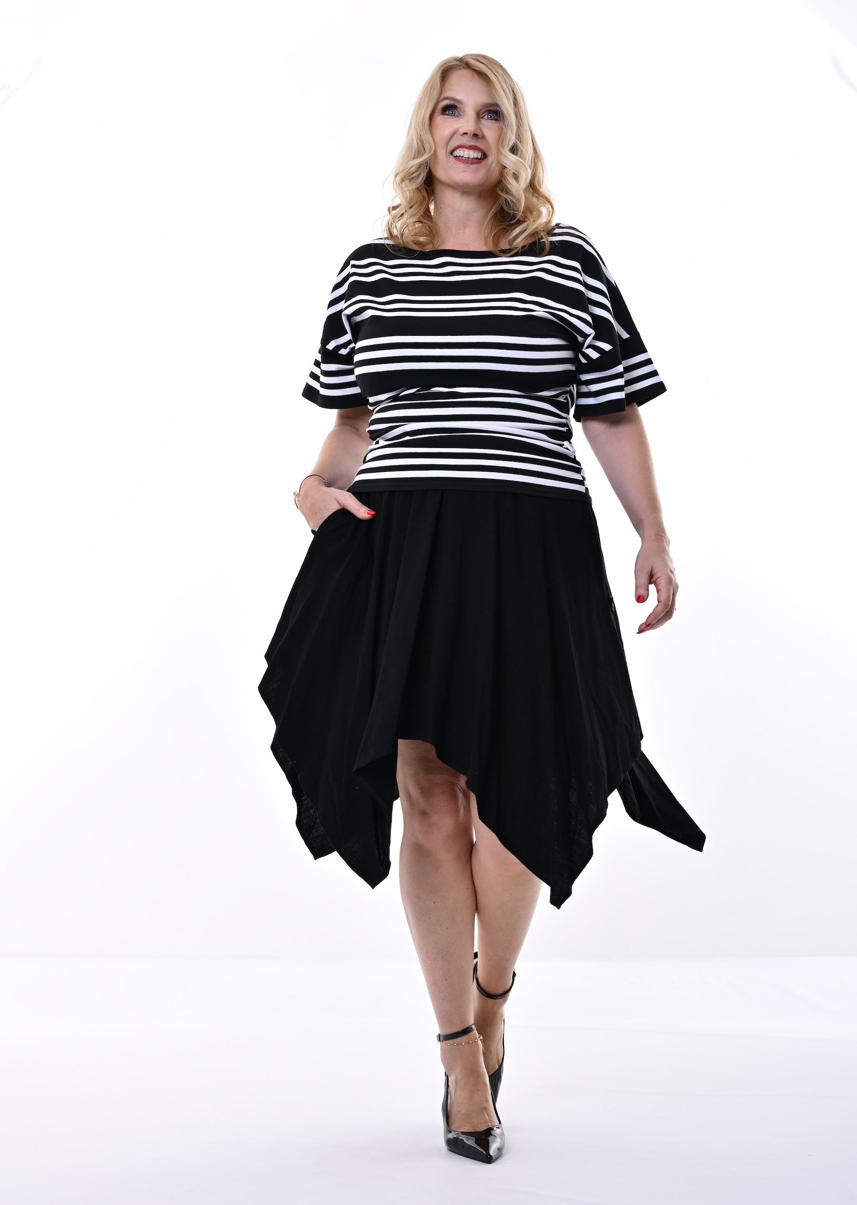 A woman modeling a black and white horizontally striped short-sleeve top with a black high-low skirt, paired with black heels, against a bright white studio background.