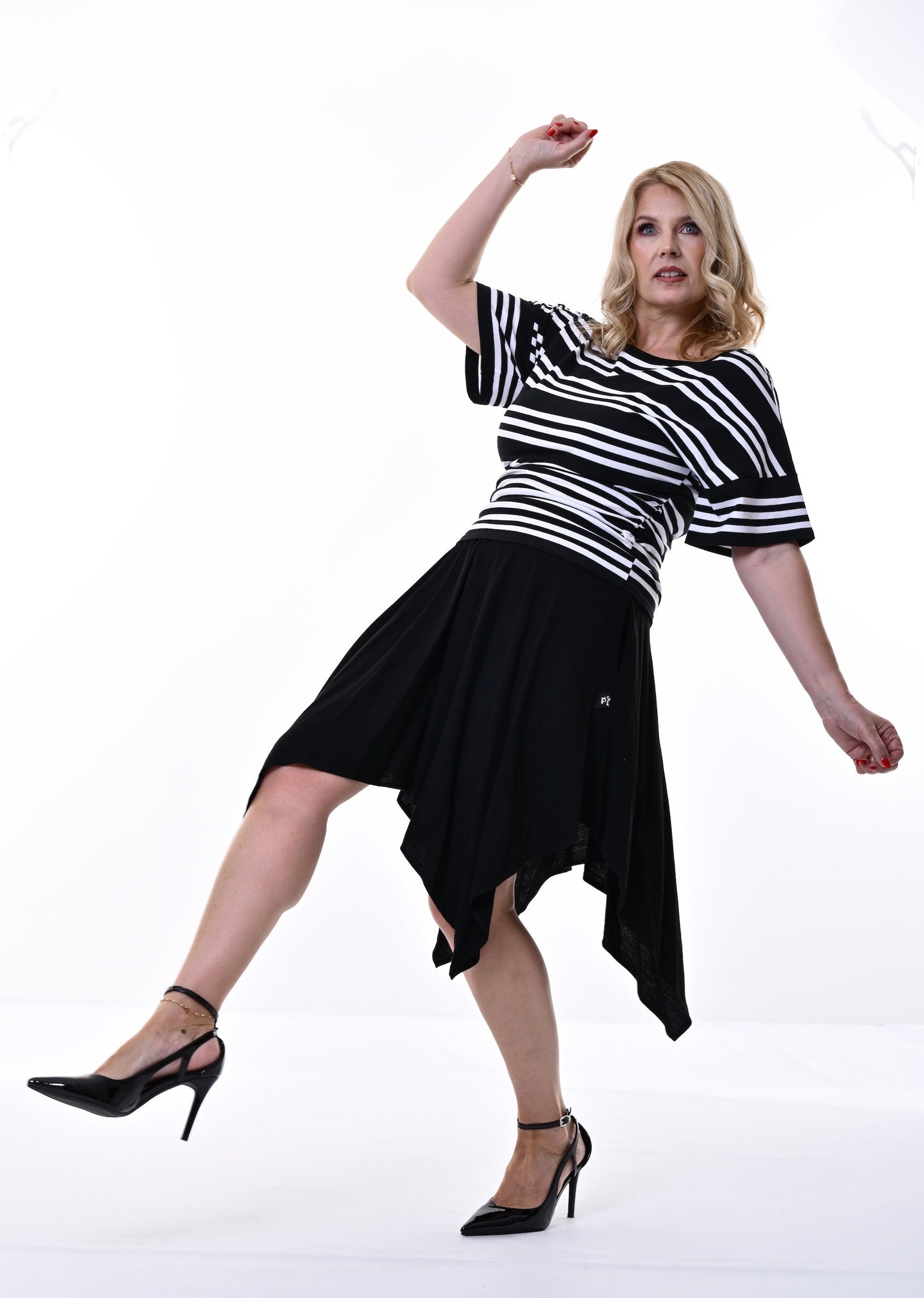 A woman posing in a black and white horizontally striped short-sleeve top with a black high-low skirt and black high heels against a white studio background.