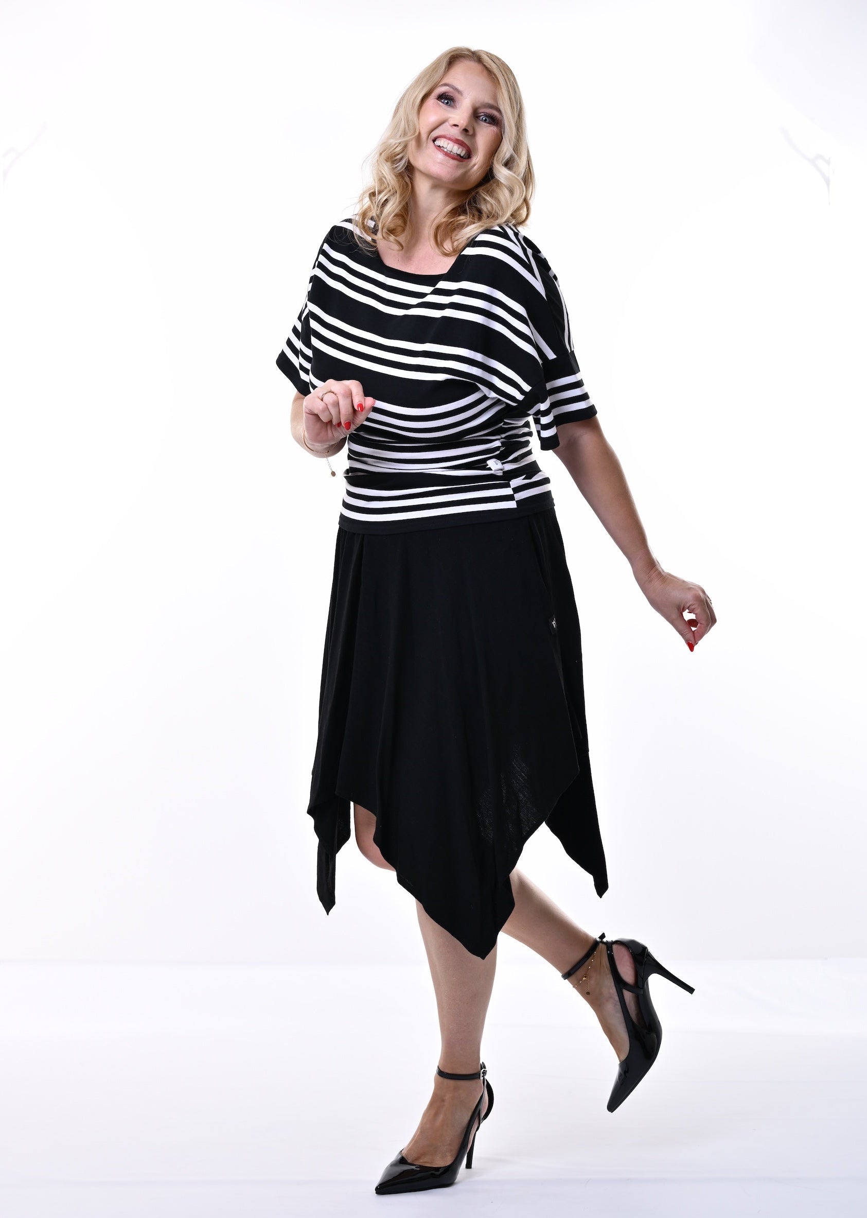 A woman modeling a black and white horizontally striped short-sleeve top with a black high-low skirt and black heels, posing in a bright white studio setting.