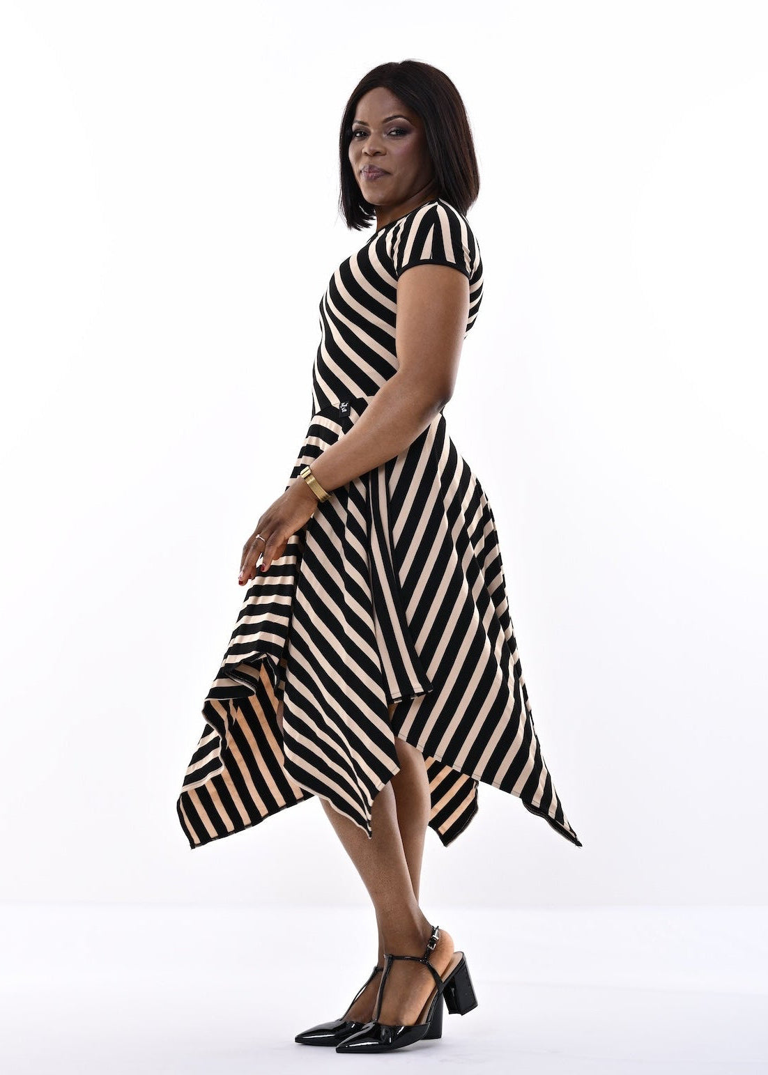 A woman modeling a black and sand diagonally striped midi dress with a hanky hemline, paired with black strappy heels, set against a bright white studio background.