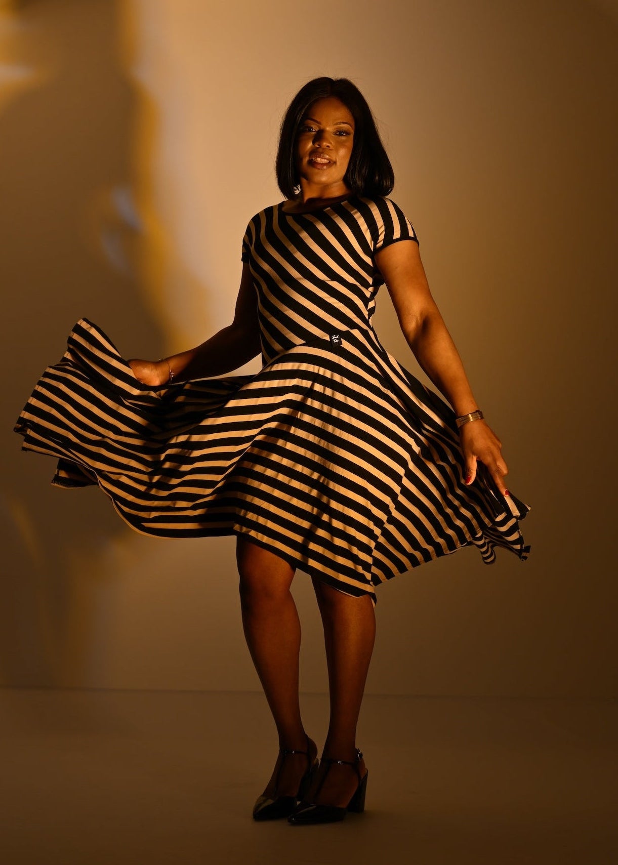 A woman wearing a black and beige striped short-sleeve dress twirls while holding out both sides of the skirt to show its volume. She is wearing black pointed-toe heels against a warm, solid beige studio background.