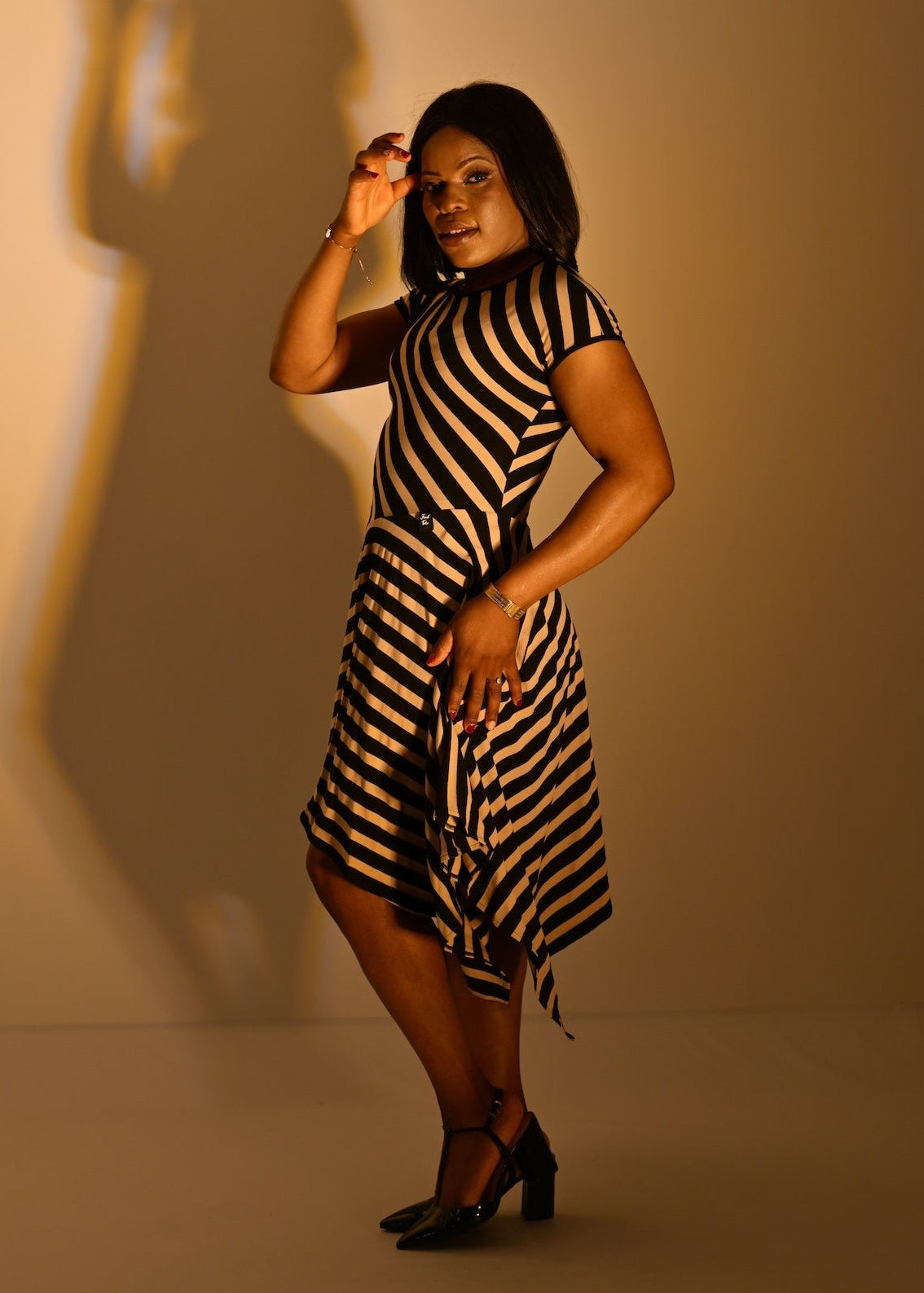 A woman modeling a short-sleeve black and sand striped hanky hem dress, standing in a studio with dramatic warm lighting and a long shadow on the wall.