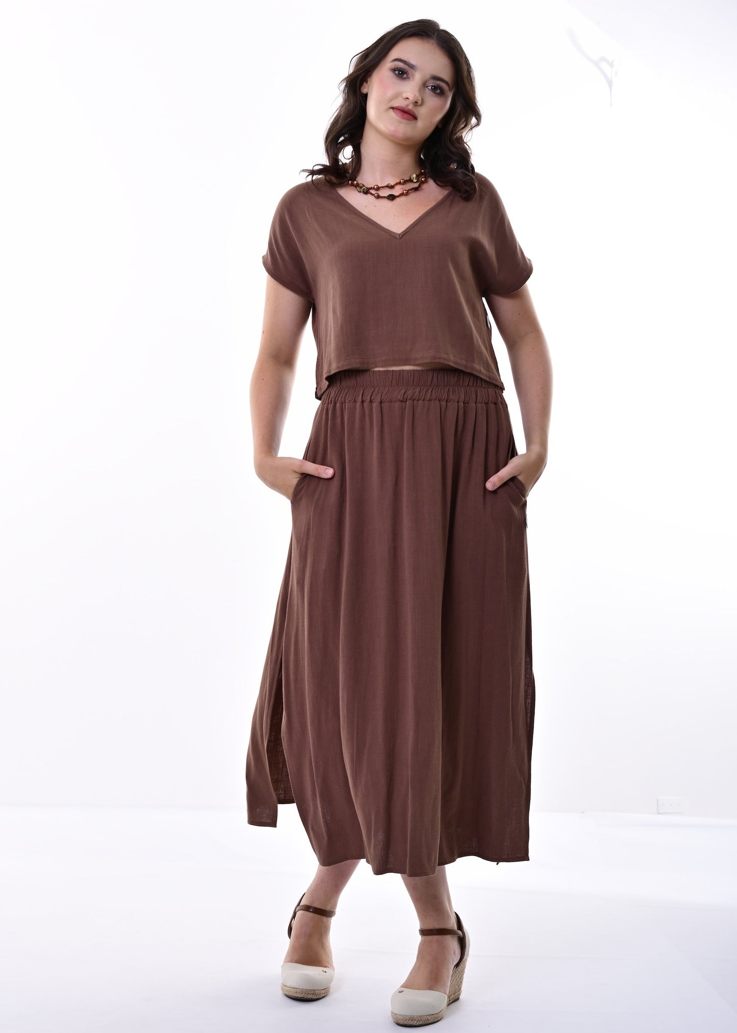 A woman modelling a brown co-ord with a short-sleeve top and a gathered skirt with side pockets, standing against a white studio background.