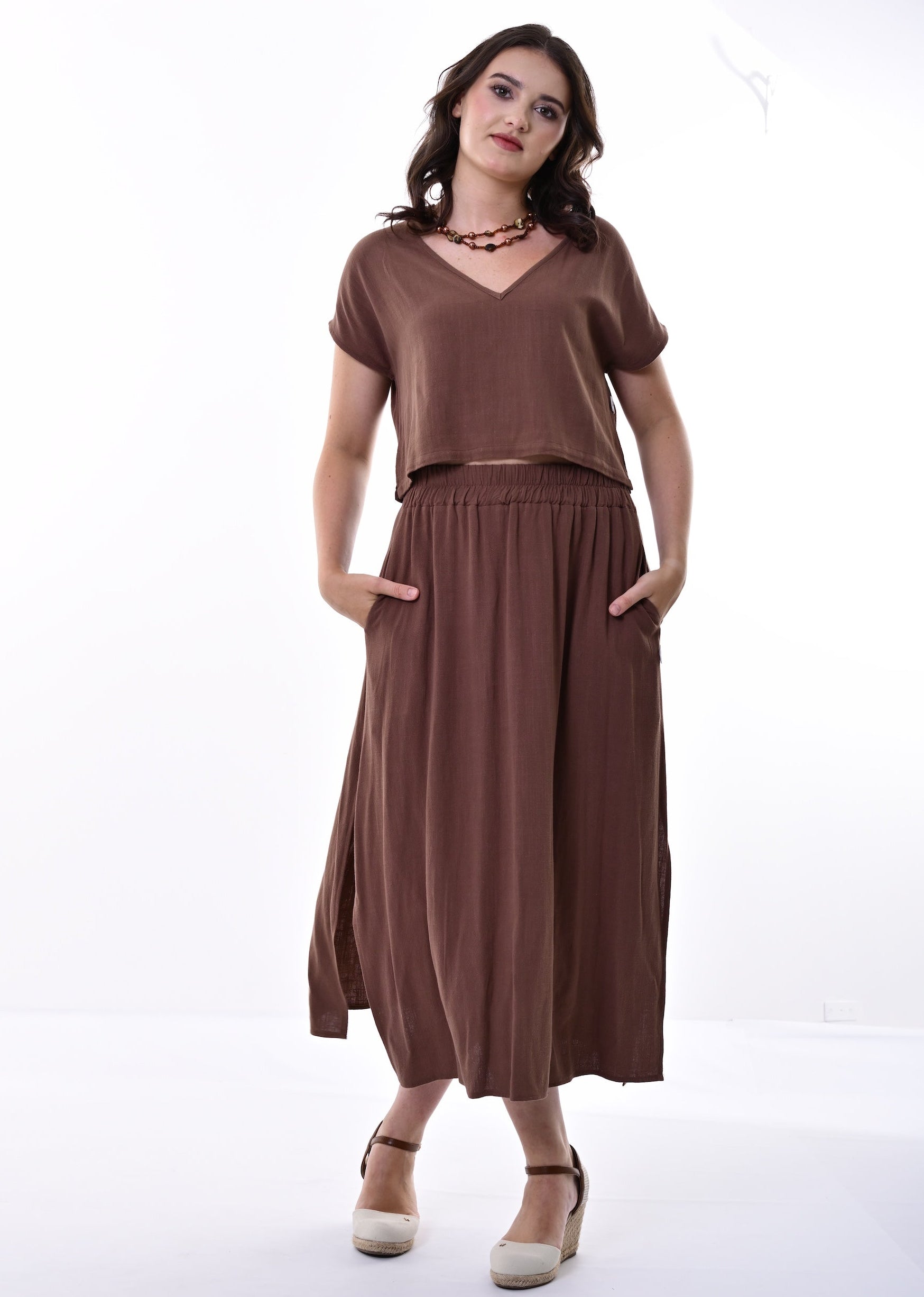 A woman modelling a brown co-ord with a short-sleeve top and a gathered skirt with side pockets, standing against a white studio background.
