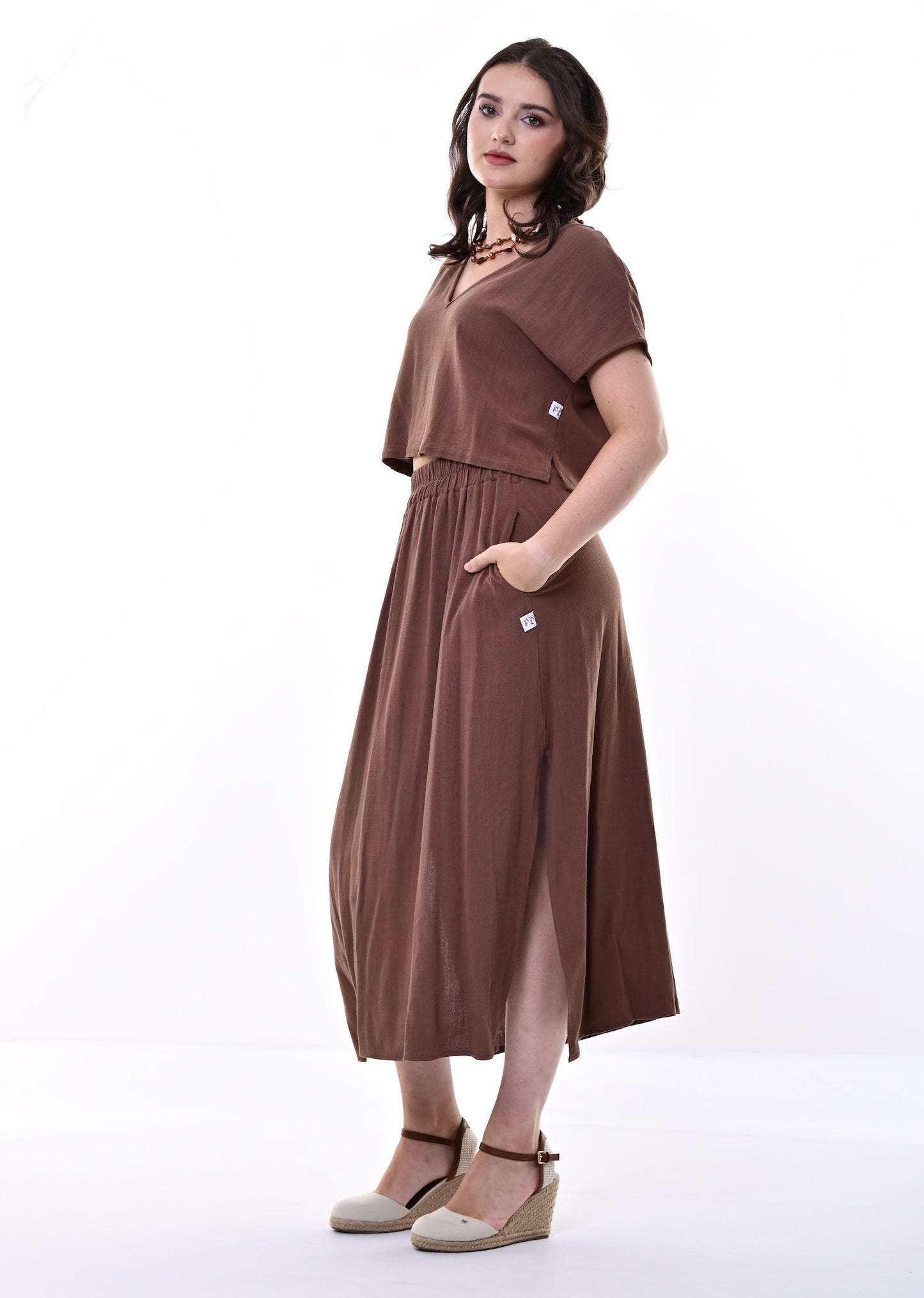 A woman modelling a brown short-sleeve top and skirt with a gathered waist and side pockets, paired with beige wedge sandals, against a plain white studio background.