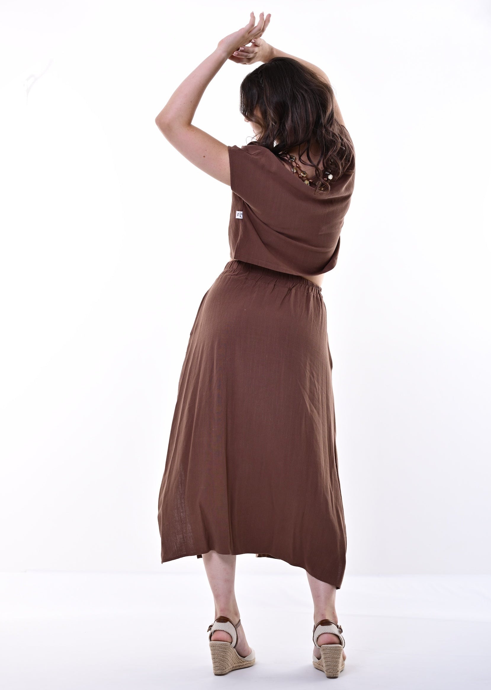 A woman wearing a brown top and skirt and beige wedge sandals, photographed from behind against a clean white studio background.