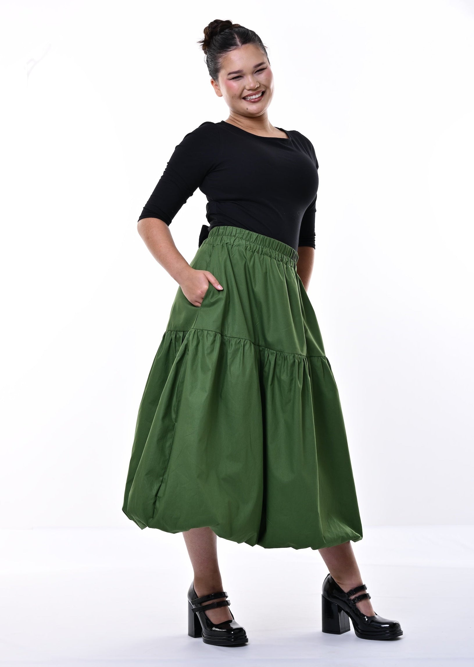 A woman modeling a tiered olive green midi skirt with side pockets, paired with a black short-sleeve top and black Mary Jane heels, against a white studio background.