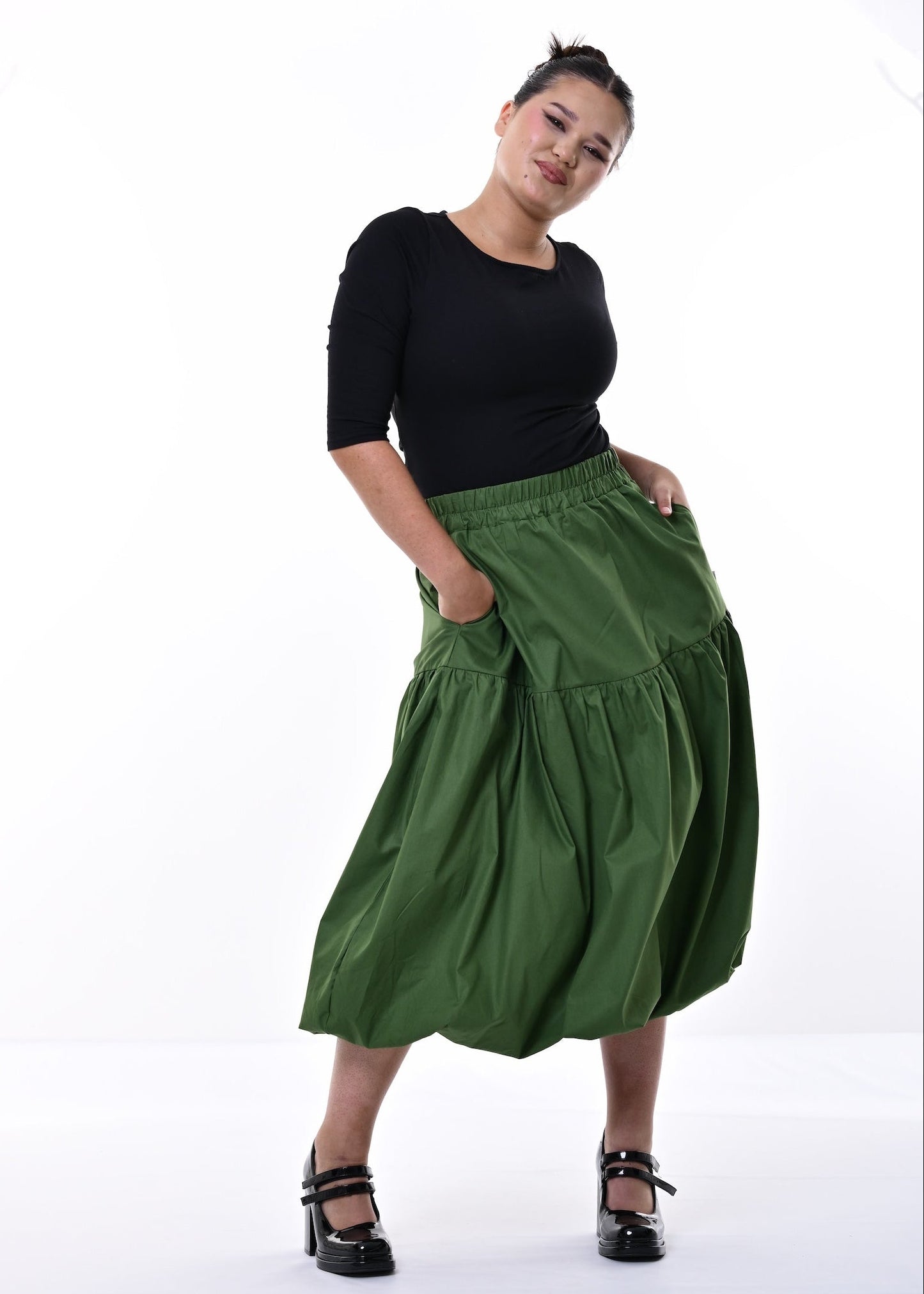 A woman modeling a black short-sleeve top and a high-waisted, tiered olive green midi skirt with side pockets, paired with black platform heels, against a white studio background.