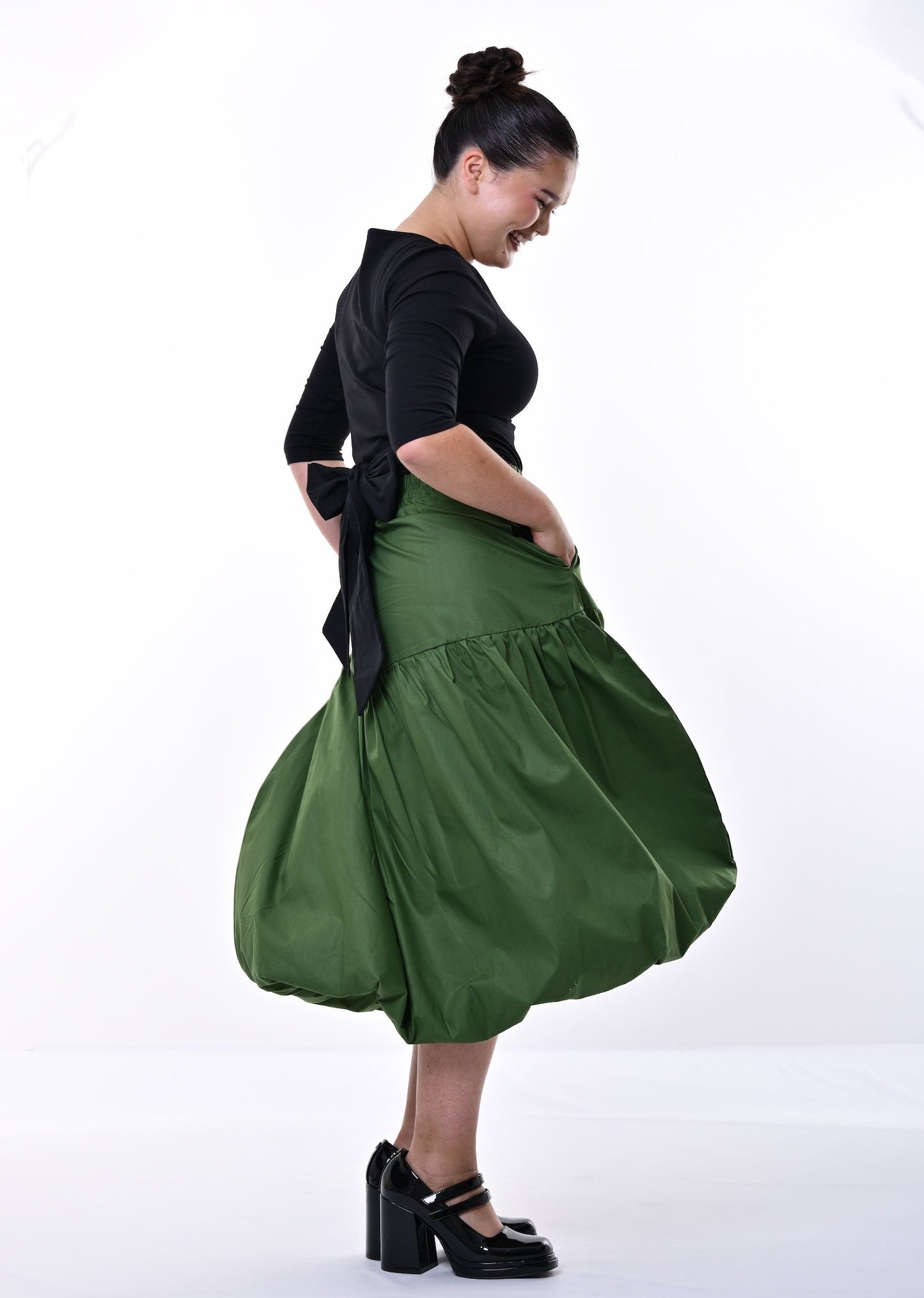 A woman modeling a black short-sleeve top and a voluminous olive green midi skirt with black platform heels, standing against a white studio background.