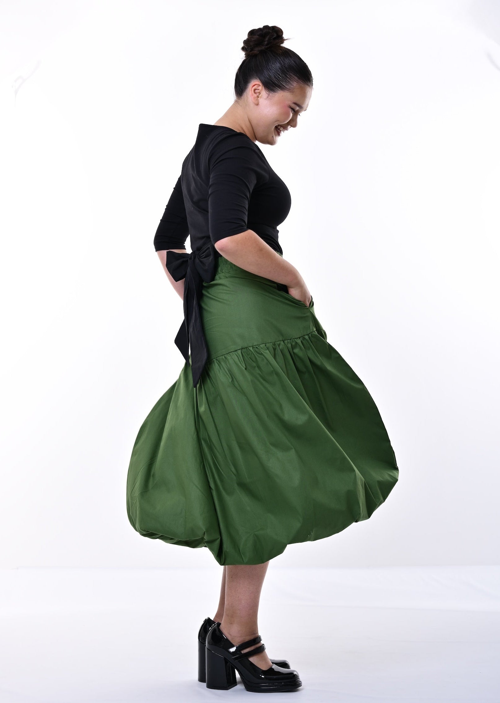 A woman modeling a black short-sleeve top and a voluminous olive green midi skirt with black platform heels, standing against a white studio background.