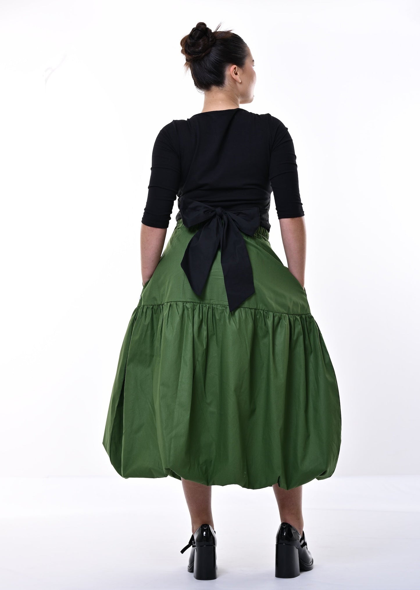 A woman viewed from behind wearing a black three-quarter sleeve top and a voluminous olive green skirt with a large black bow at the back waist, standing against a white studio background.