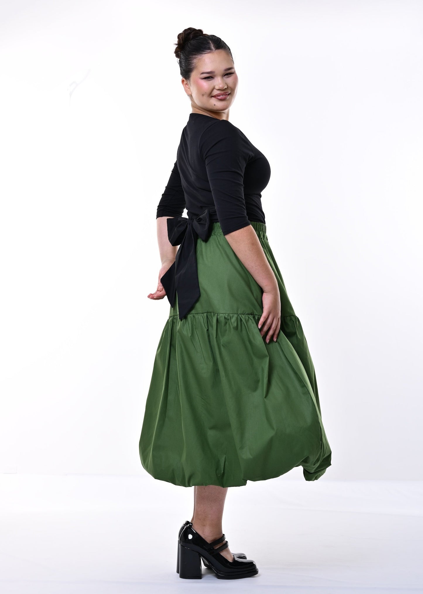 A woman modeling a two-piece outfit consisting of a black short-sleeve top and a voluminous olive green tiered skirt, paired with black Mary Jane heels, set against a plain white studio background.