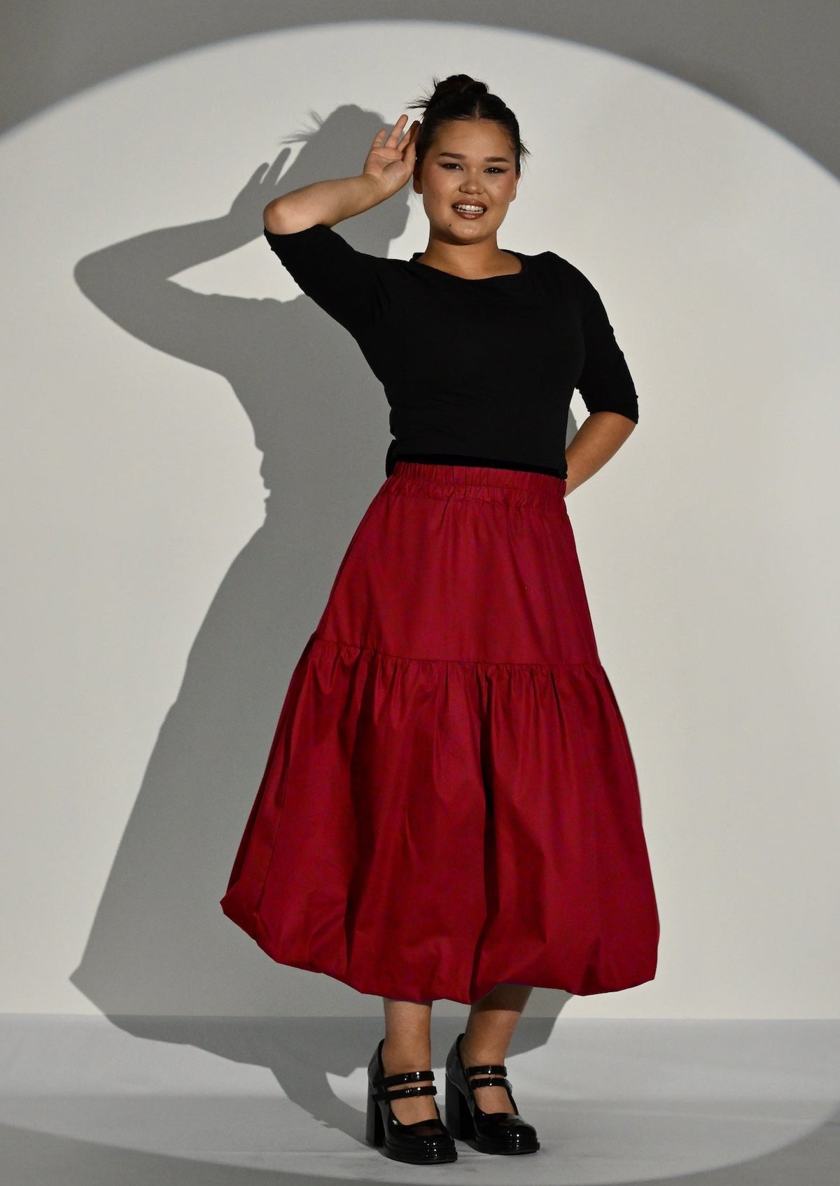 A woman posing in a black 3/4 sleeve top and a tiered red puffball skirt against a white studio backdrop with a spotlight effect.