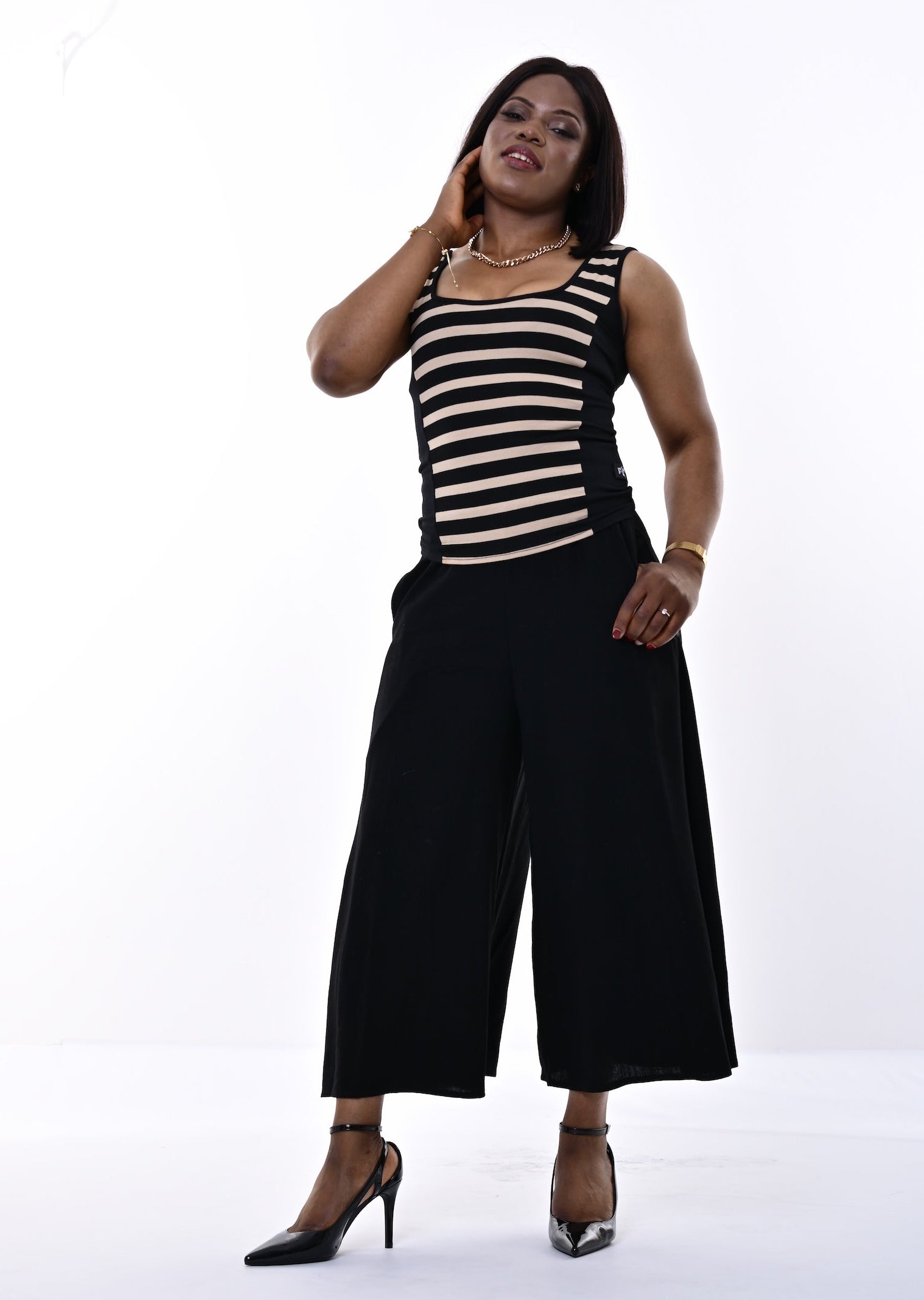 A woman modeling a sleeveless black and beige striped top paired with wide-leg black pants and black high heels, standing against a plain white studio background.