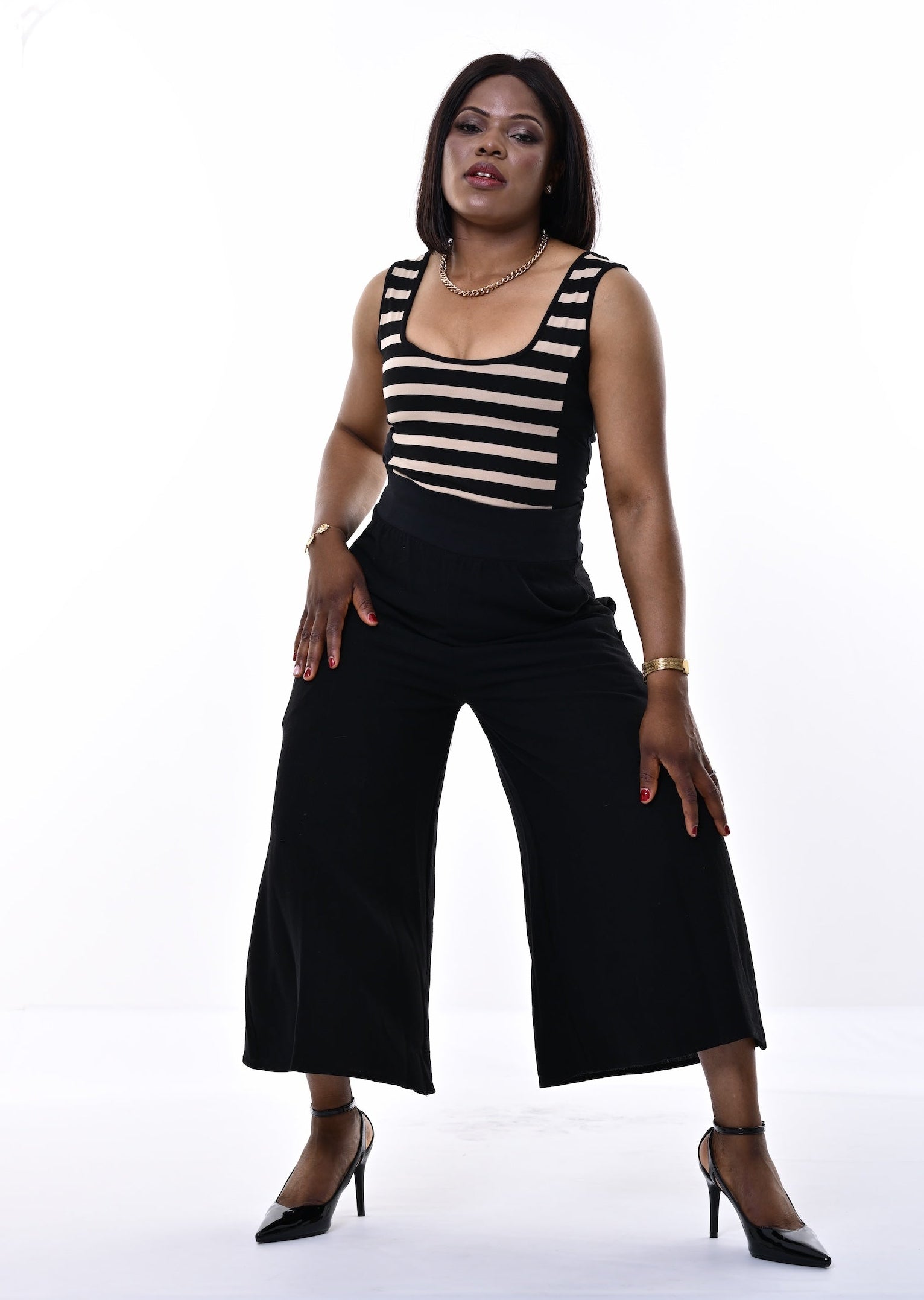 A woman modeling a sleeveless black and white horizontally striped top paired with black wide-leg cropped pants and black high heels, standing against a bright white studio background.