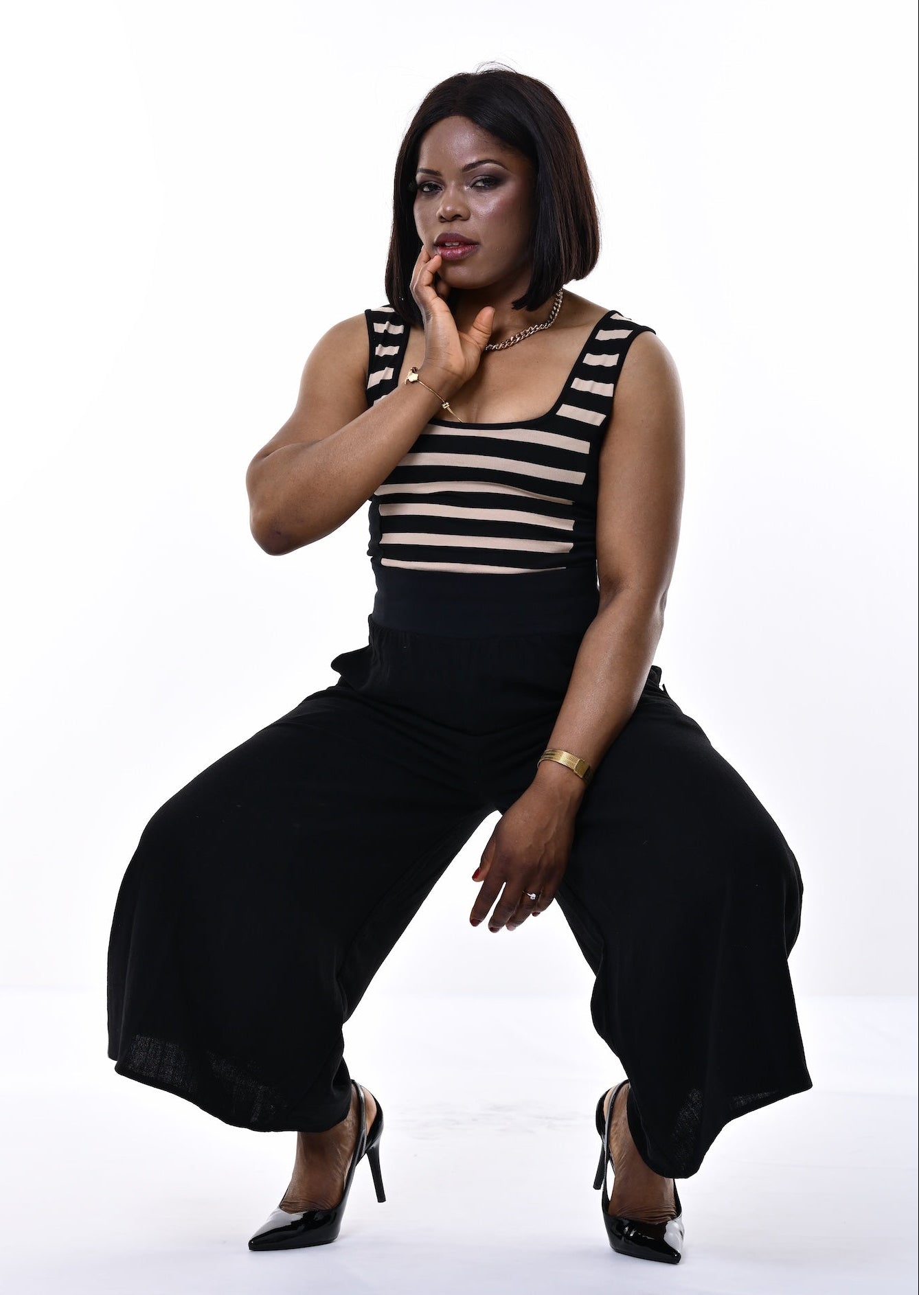 A woman posing in a black sleeveless jumpsuit with horizontal beige stripes on the chest and wide-leg pants, set against a plain white studio background.