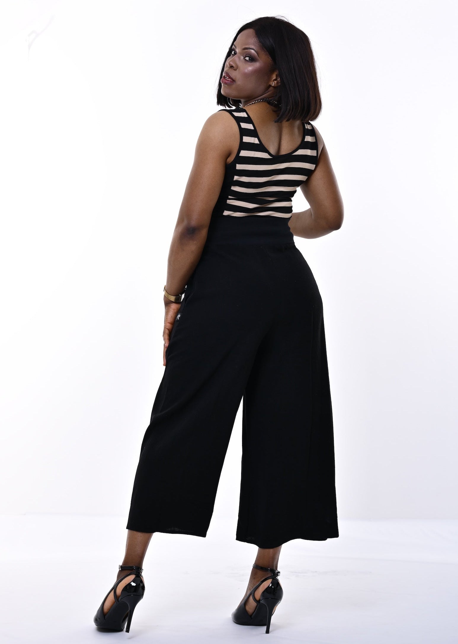 A woman posing in a studio setting, wearing a black and sand horizontally striped sleeveless top paired with high-waisted black wide-leg cropped pants and black strappy heels.