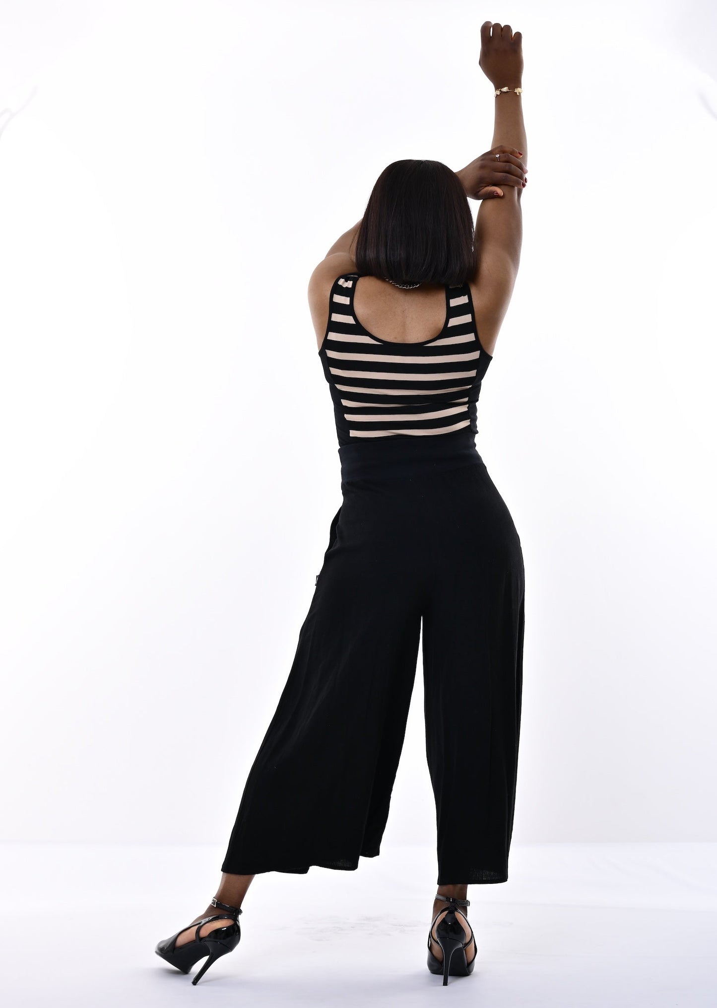 A woman posing from behind in a studio setting, wearing a black and sand horizontally striped sleeveless top and black wide-leg cropped pants with black high heels.