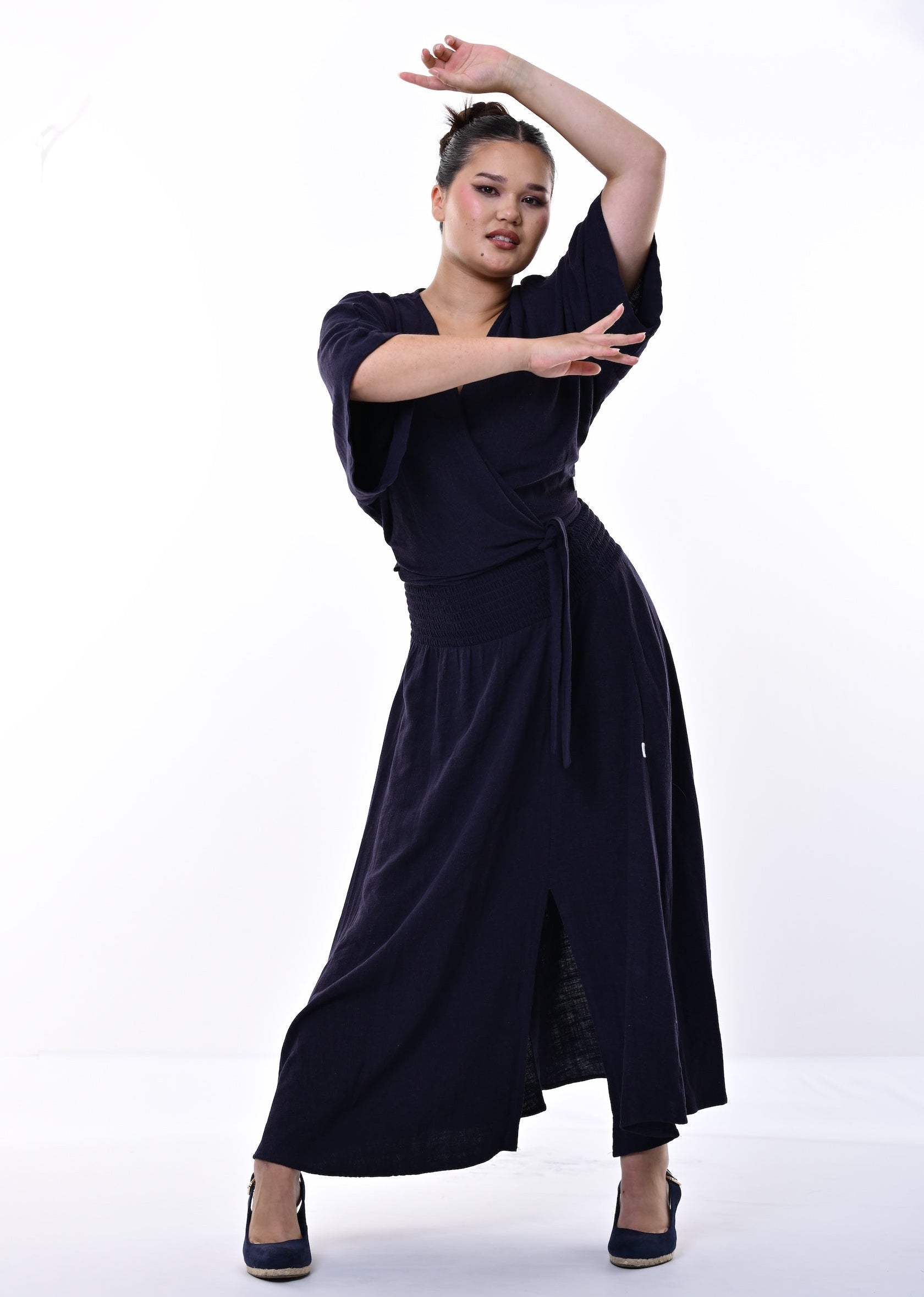 A woman modelling a dark navy blue wrap-style top and skirt with short wide sleeves and a side slit, posing against a bright white studio background.
