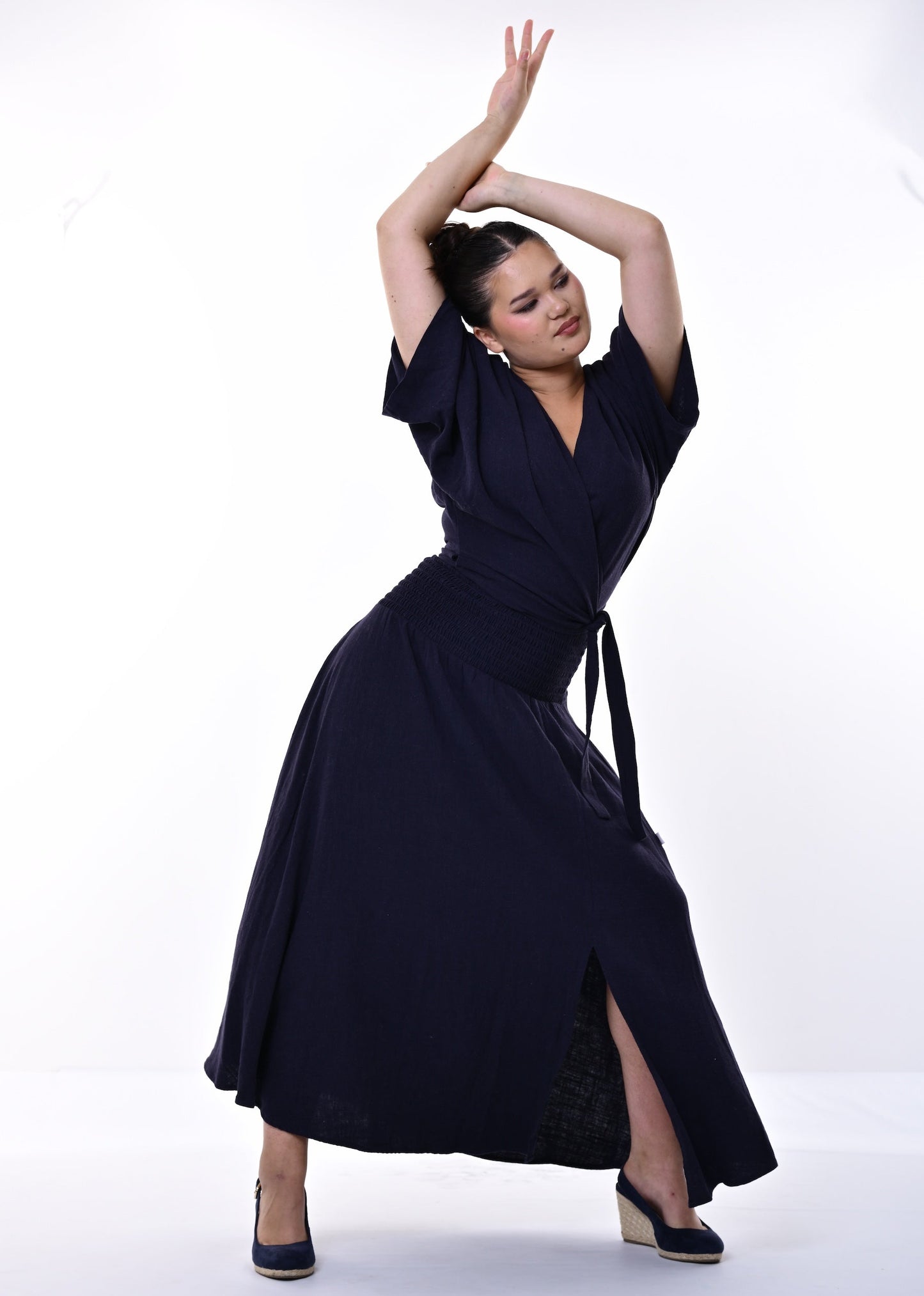 A woman modelling a dark navy blue wrap-style top and skirt dress with short sleeves, a tie waist, and a side slit, posing against a plain white studio background.