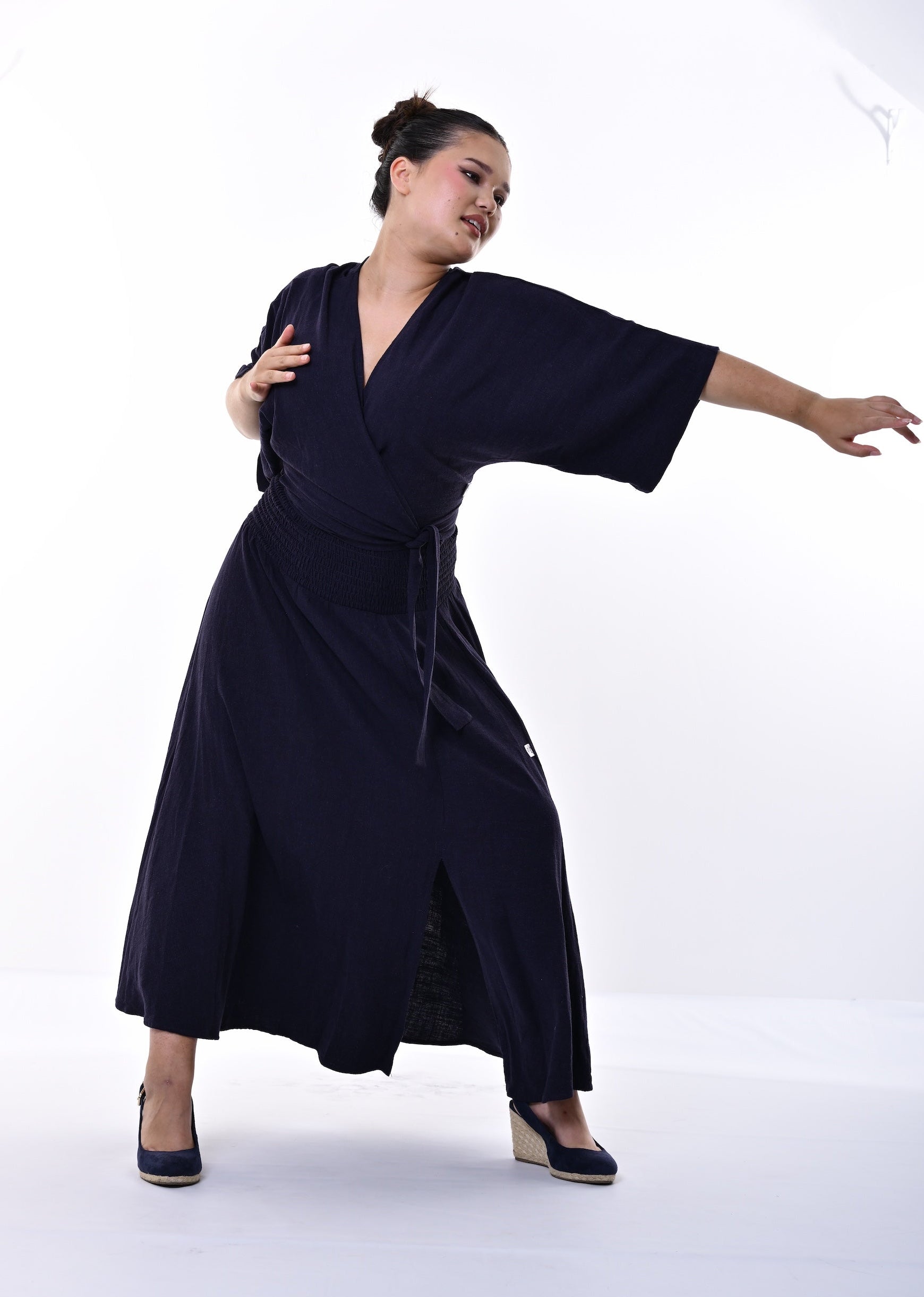 A woman modelling a dark navy wrap-style top and skirt with wide sleeves and a tie waist, posed against a plain white studio background.
