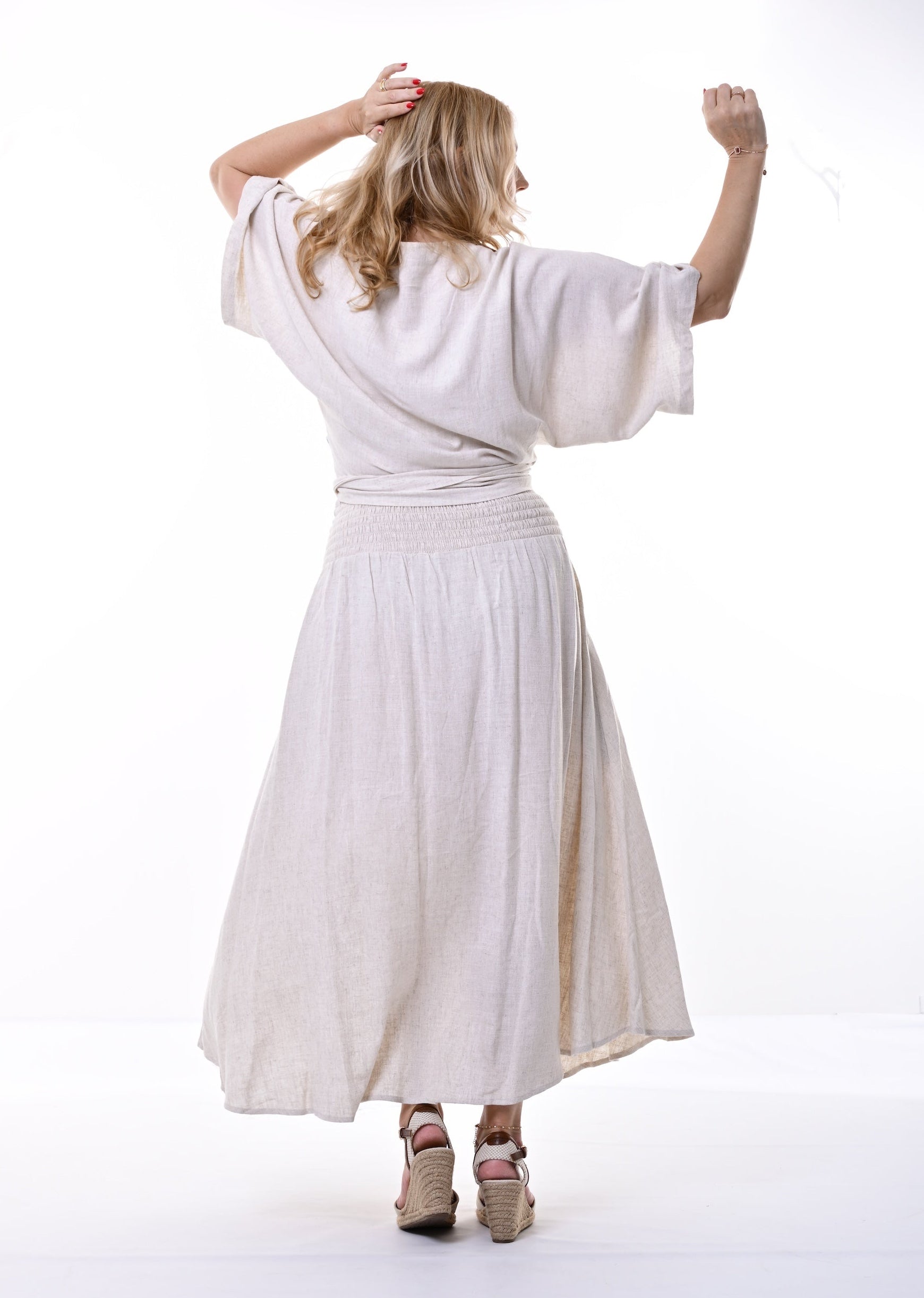 A woman viewed from behind modelling an oatmeal linen wrap top with wide sleeves and a matching skirt with leg slit, paired with beige high heels, against a bright white studio background.