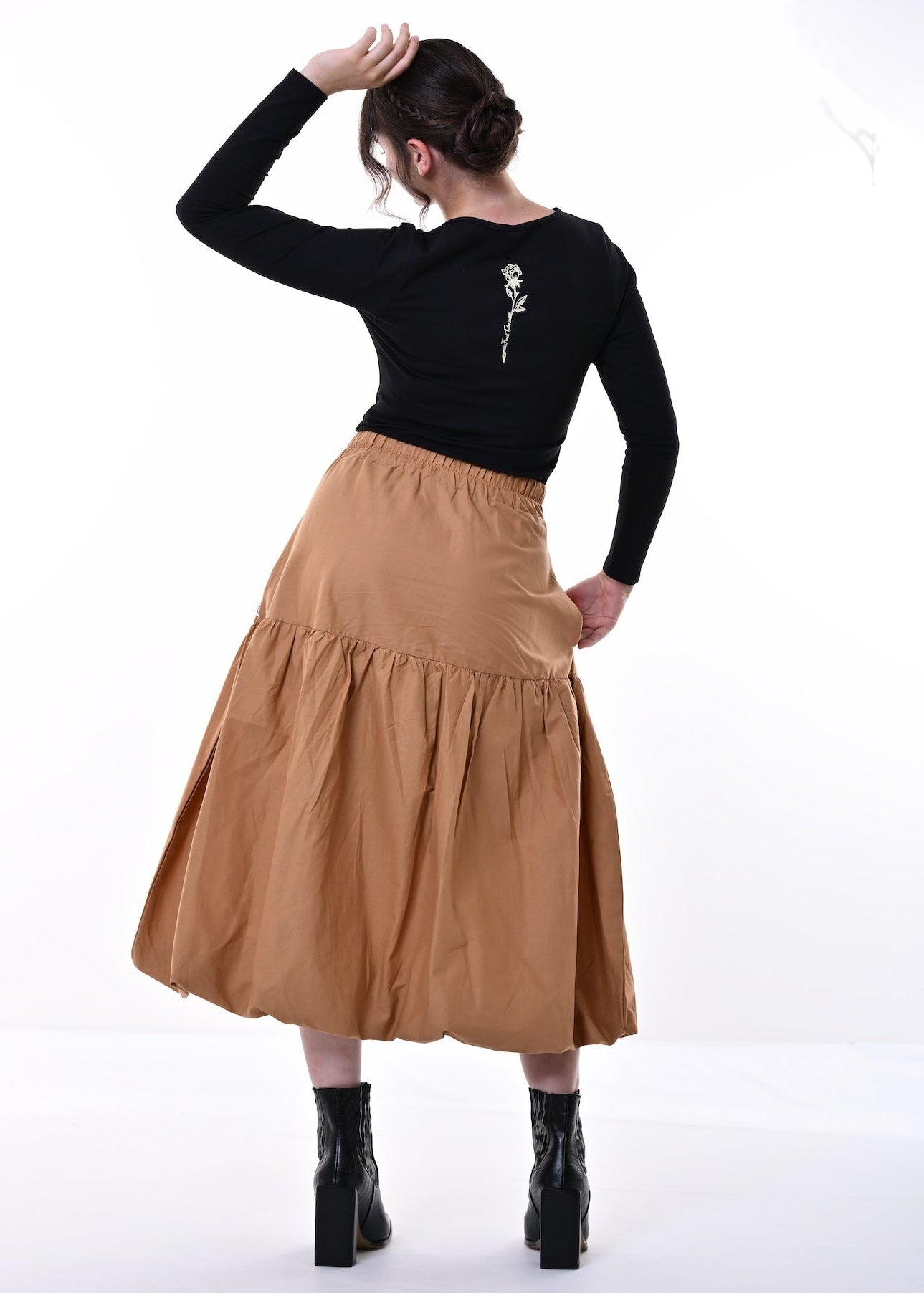A woman is shown from behind wearing a black long-sleeve top with a cream rose graphic on the back and a tiered tan skirt, paired with black ankle boots, against a white studio background.