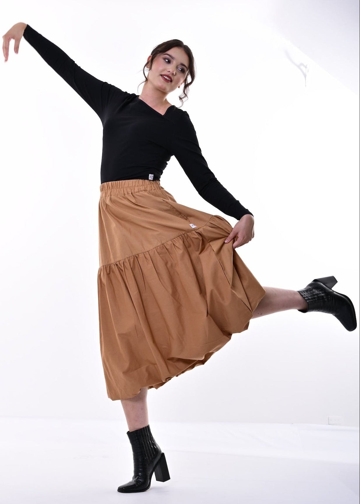 A woman posing in a studio wearing a black long sleeved asymmetrical top and a tiered tan midi skirt, paired with black ankle boots.