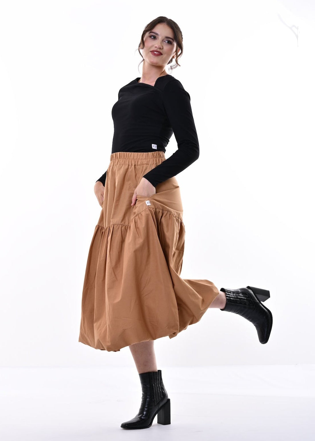 A woman modeling a high-waisted, tiered tan midi skirt paired with a black long-sleeve top and black ankle boots, posing in a bright white studio setting.