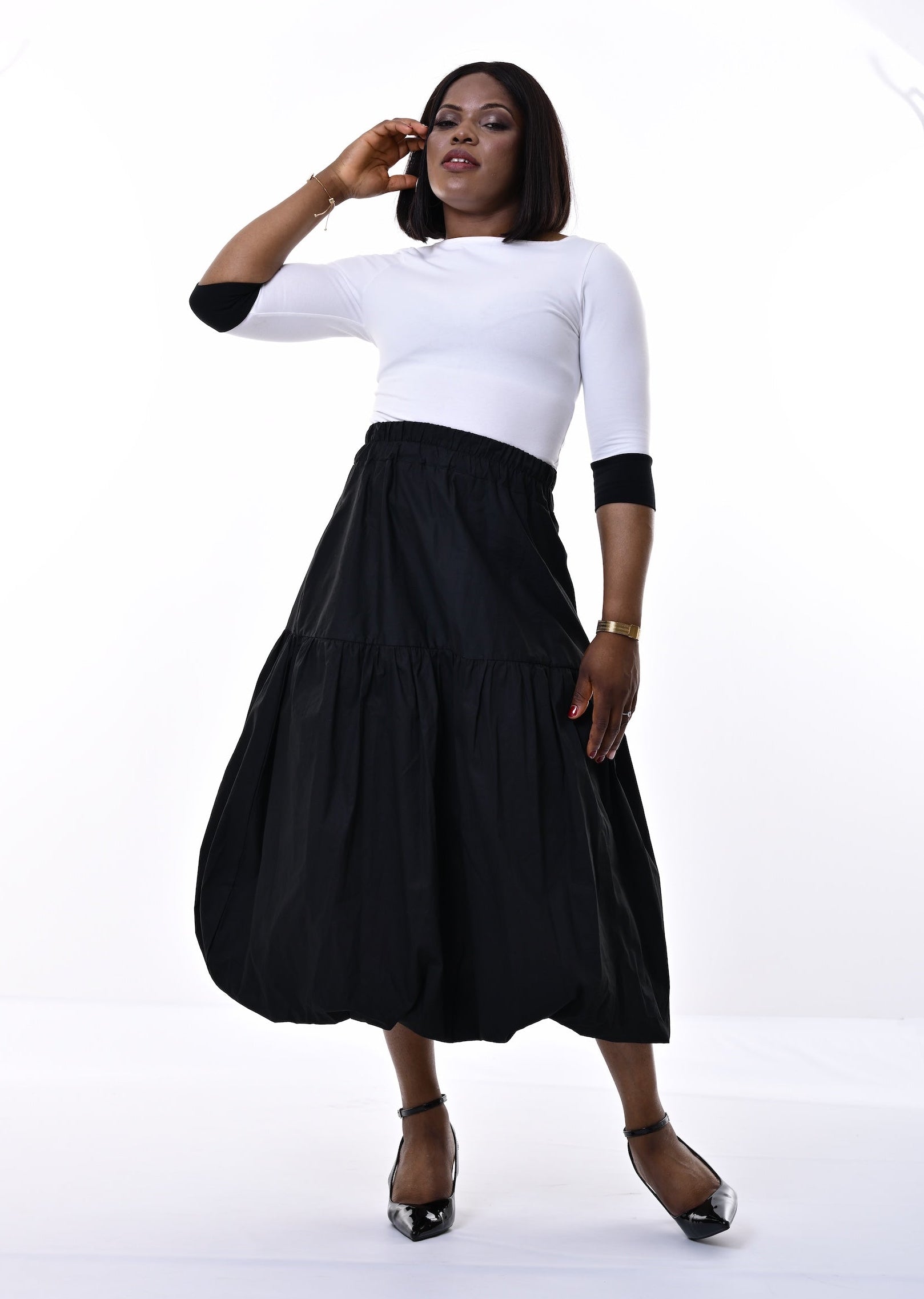 A woman posing in a white mock-neck top with three-quarter black sleeves and a high-waisted, tiered black midi skirt, paired with black heels, against a bright white studio background.