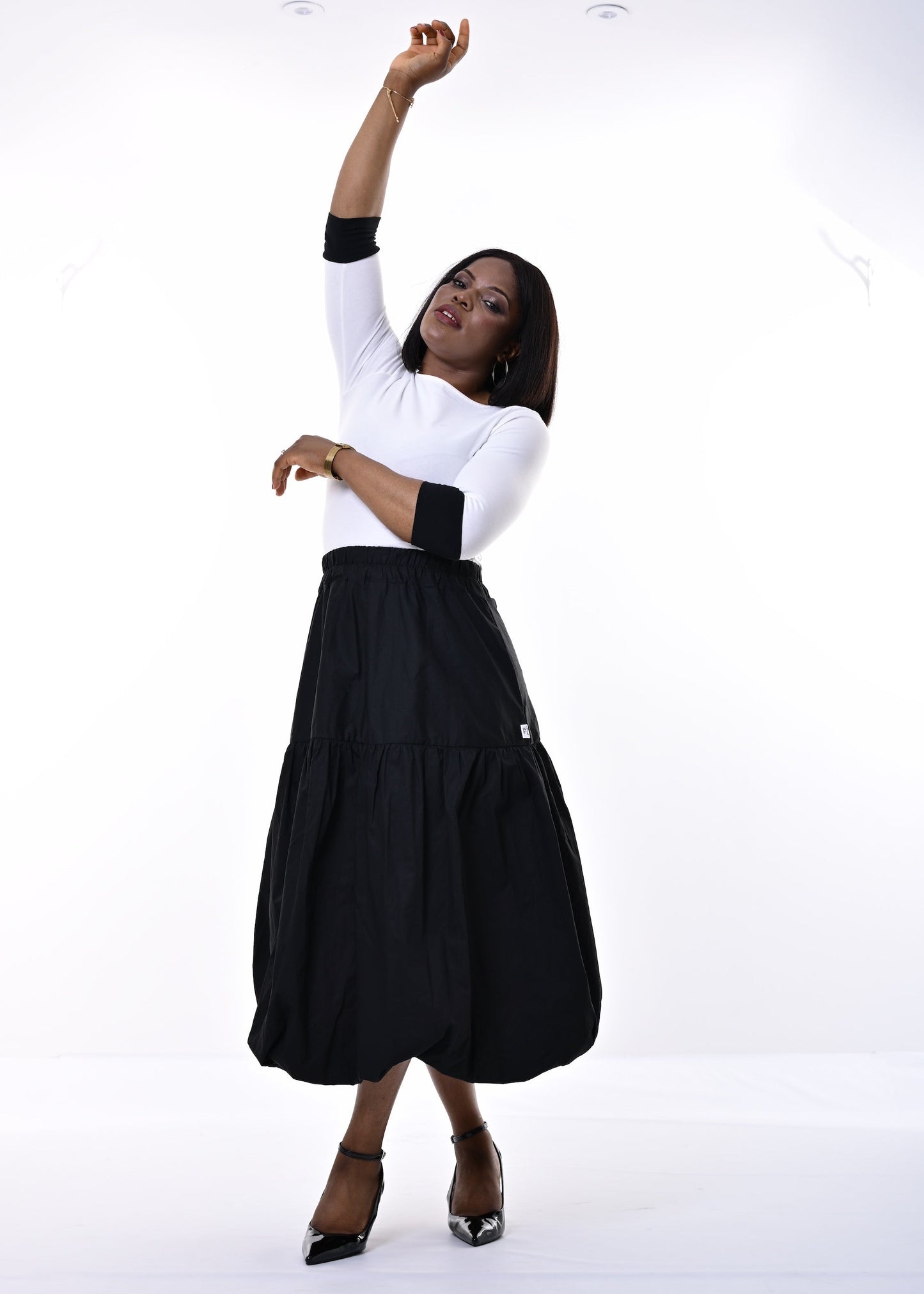 A woman modeling a white long-sleeve top with black elbow-length sleeves and a black tiered midi skirt, paired with black pointed-toe heels, against a bright white studio background.
