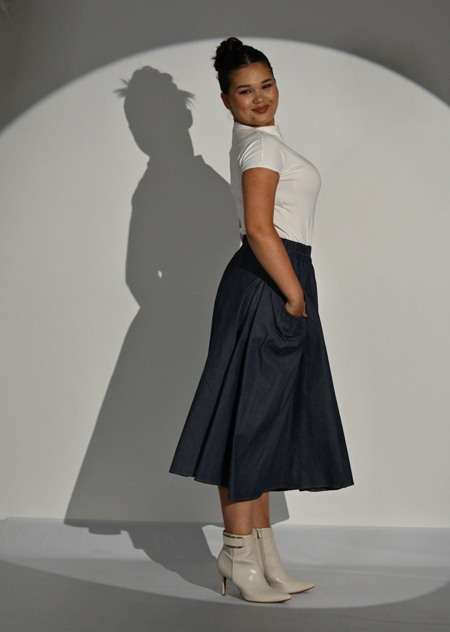 A woman stands in profile wearing a cream short-sleeve top and a dark navy midi skirt, posed inside a large spotlight on a gray studio background that casts a long shadow of her silhouette.