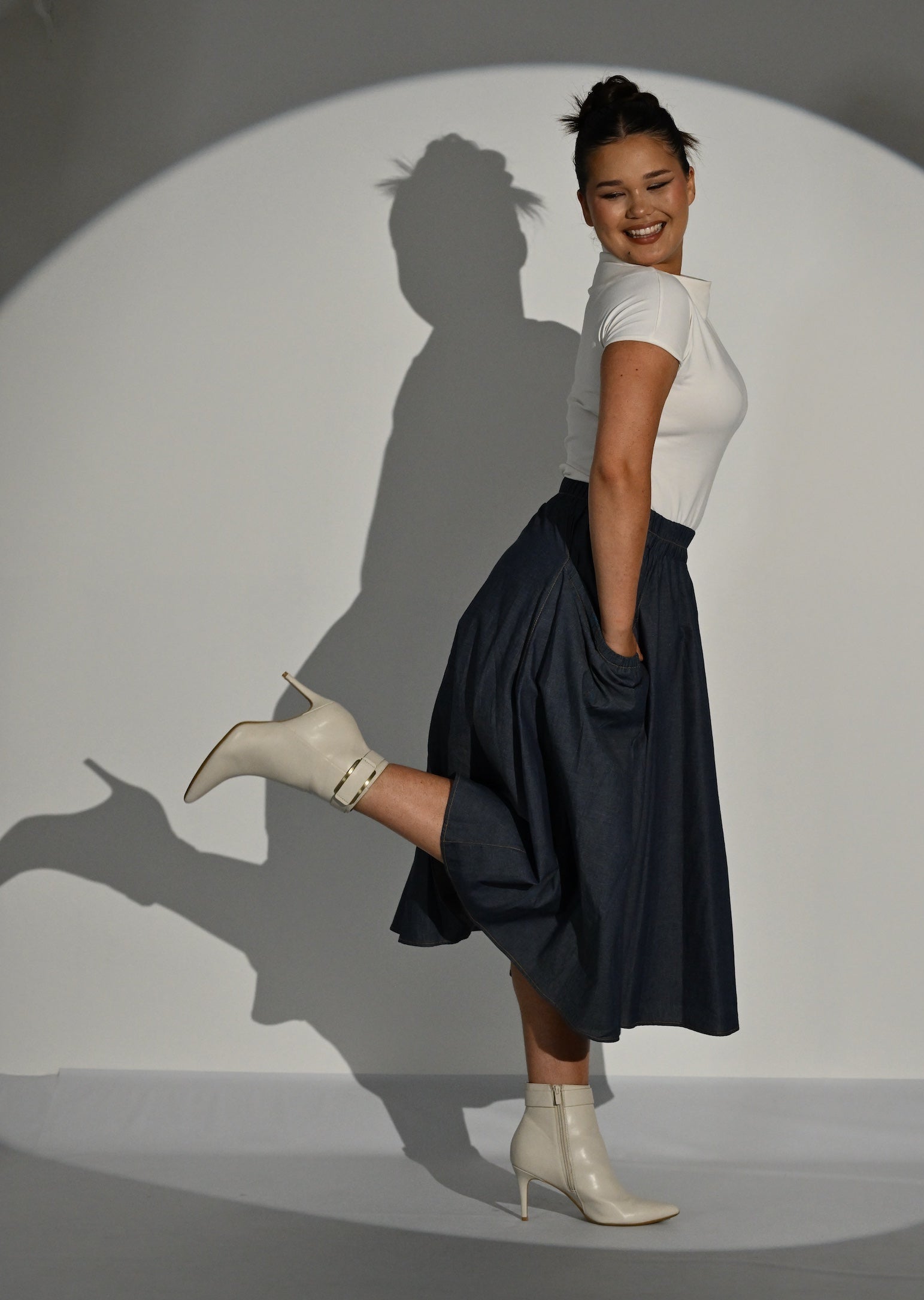 A woman posing in a studio setting under a spotlight, wearing a cream short-sleeve top, a dark navy midi skirt, and white ankle boots.