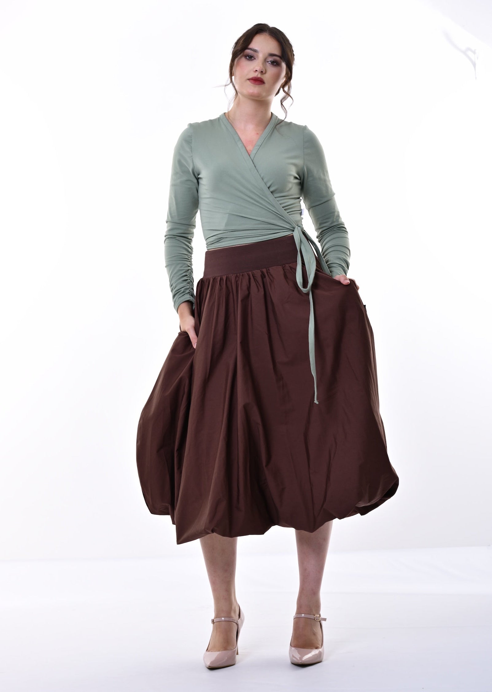 A woman modeling a sage green long-sleeve wrap top and a dark brown pleated midi skirt with a matching tie belt, standing against a white studio background.