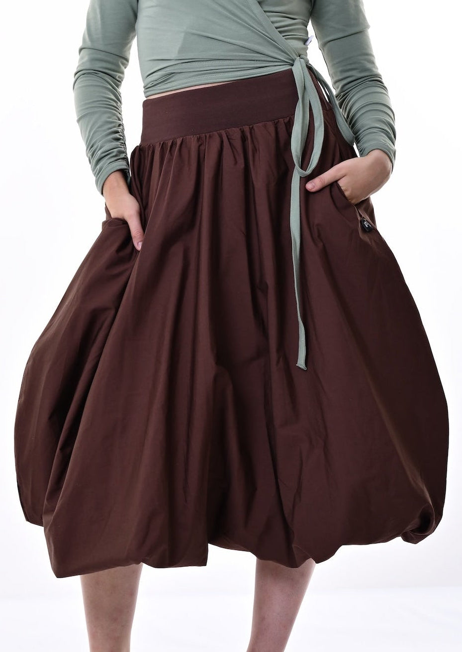 A model stands against a bright white studio background wearing a sage green long-sleeve wrap top and a voluminous dark brown pleated midi skirt with side pockets.