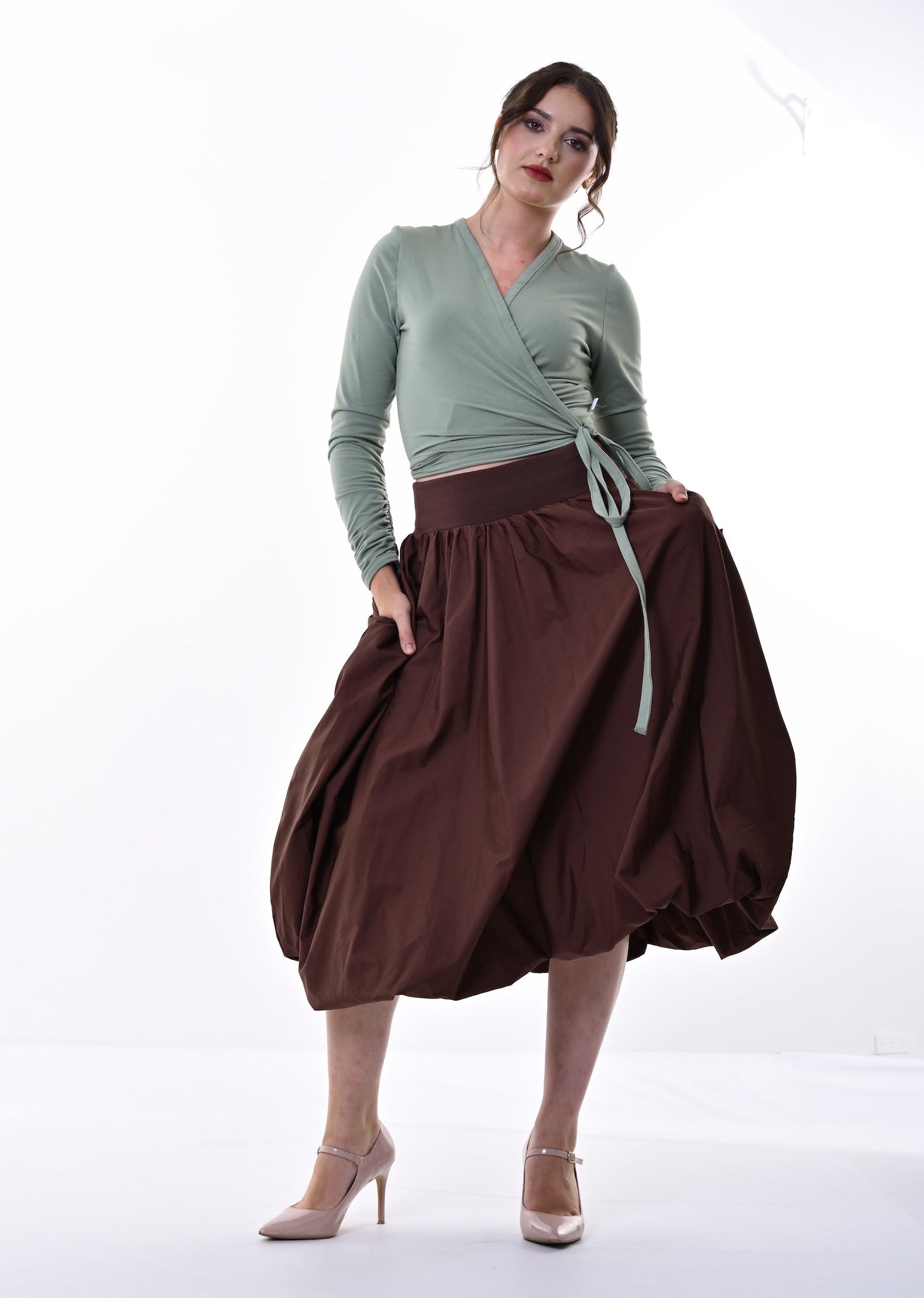 A woman modeling a sage green wrap top and a dark brown pleated midi skirt with side pockets, standing against a bright white studio background.