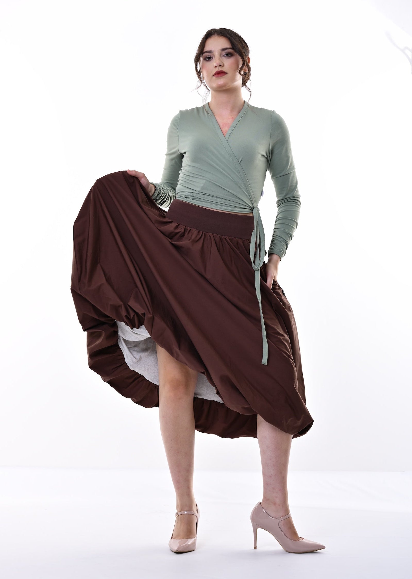 A woman modeling a sage green long-sleeve wrap top and a flowing dark brown skirt, holding up the skirt to show its volume against a bright white studio background.