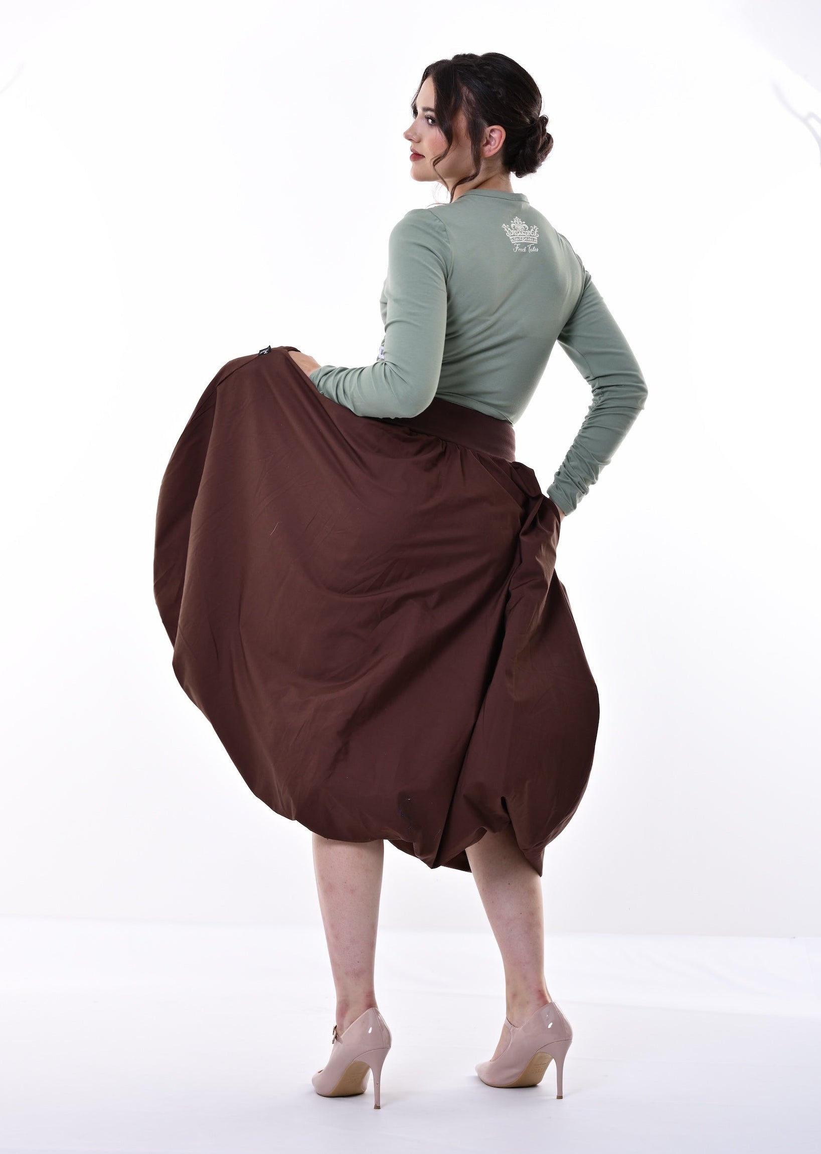 A woman posing from behind in a studio, wearing a sage green long-sleeve top and a voluminous brown skirt, holding the skirt out to show its drape and volume. She is wearing beige high heels against a bright white background.