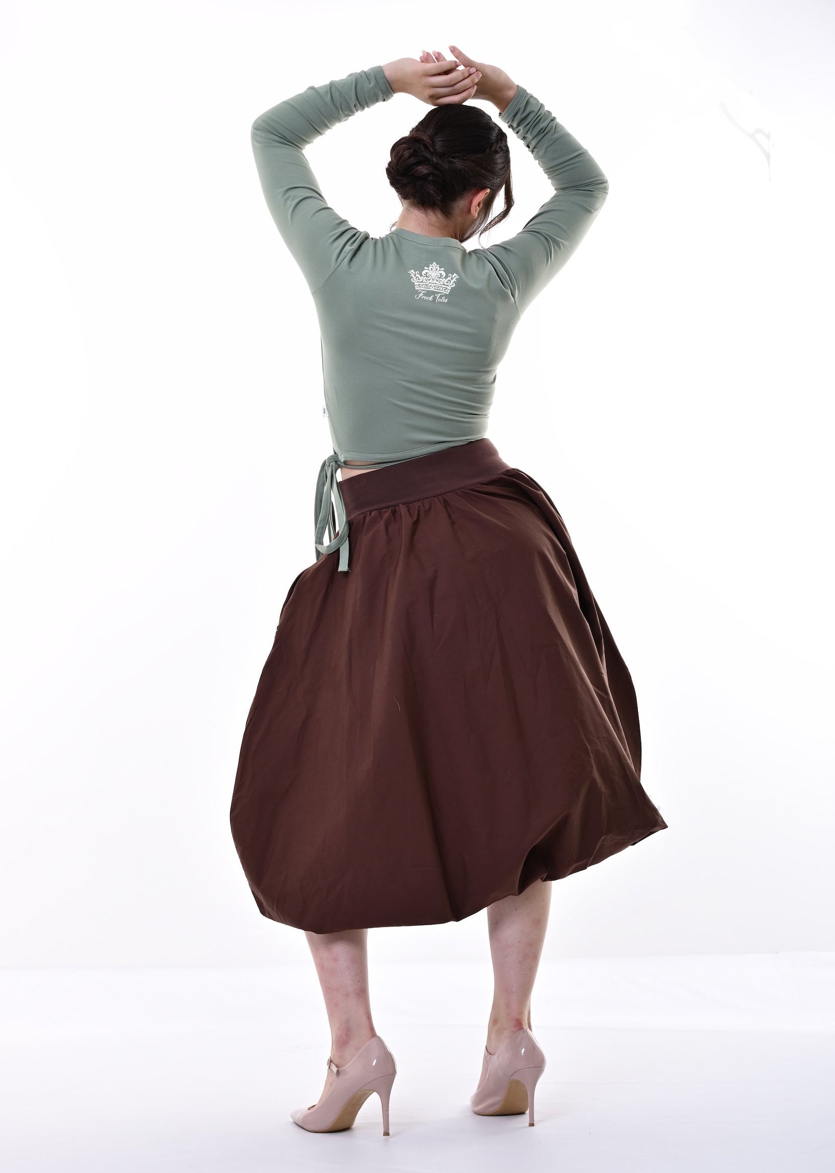A woman viewed from behind wearing a sage green long-sleeve top and a brown high-waisted pleated skirt, posing with her hands behind her head in a bright white studio setting.