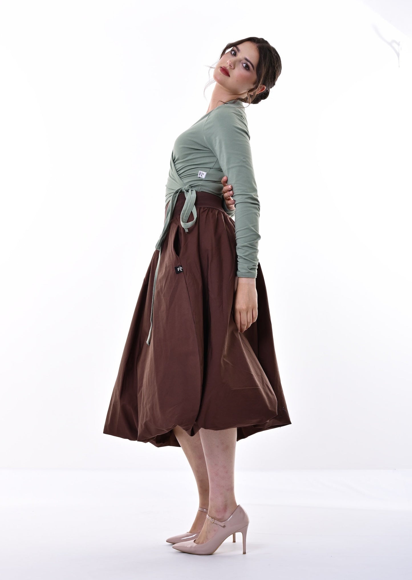 A woman posing in a studio setting, wearing a sage green long-sleeve crop top, a brown midi skirt with a tie waist, and nude high heels against a bright white background.