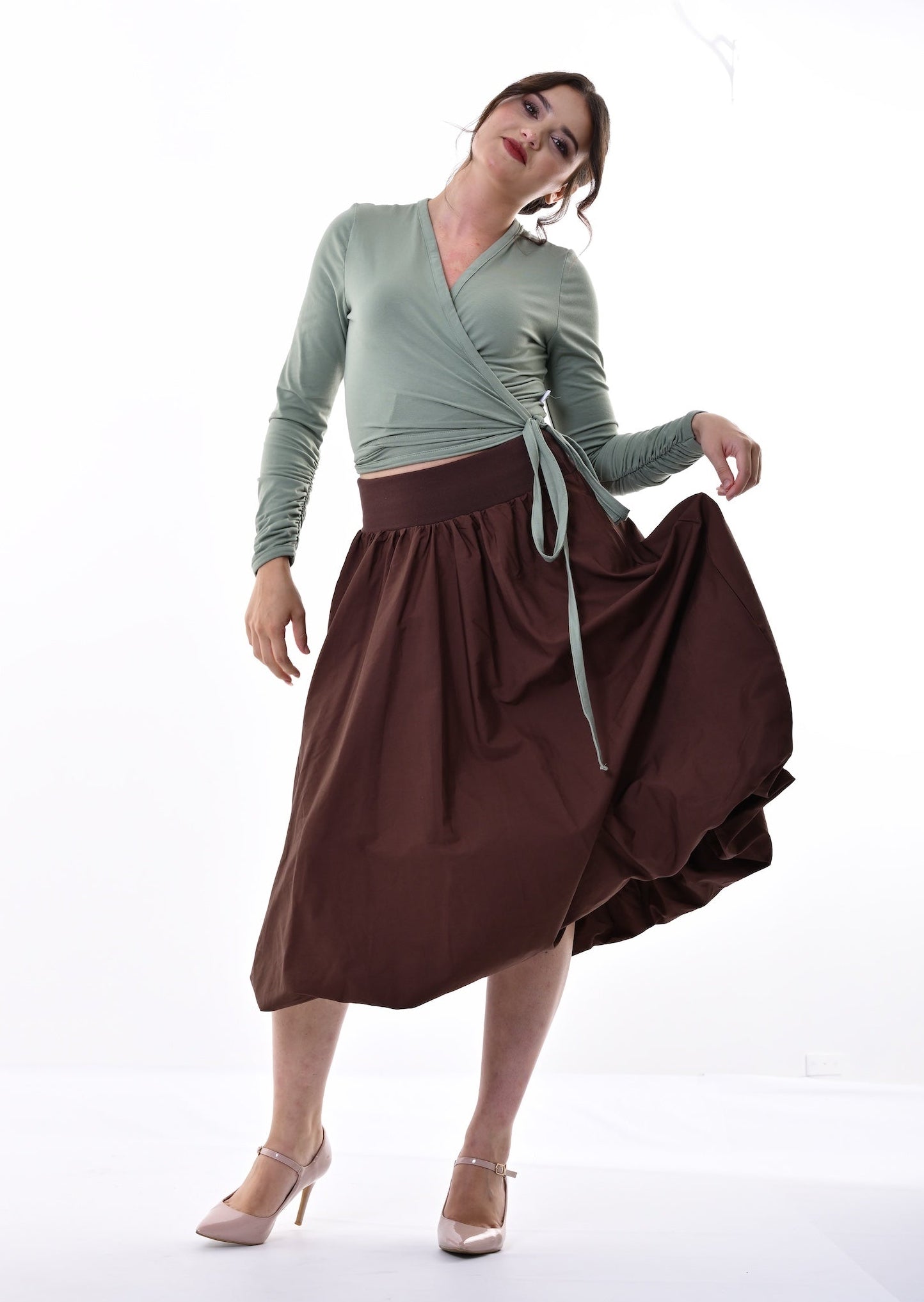 A woman modeling a sage green long-sleeve wrap top and a dark brown midi skirt with a tie waist, standing in a white studio.