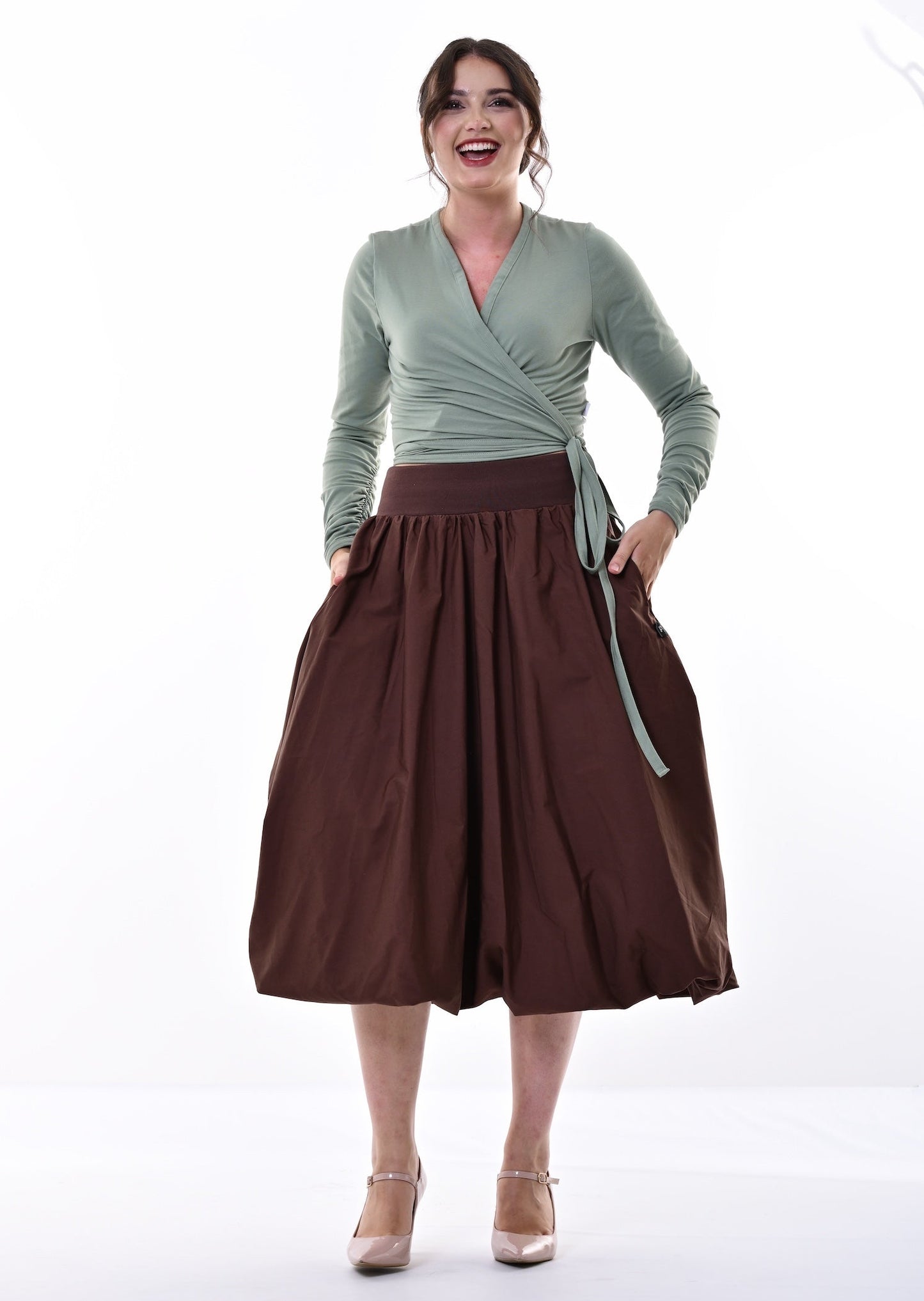 A woman modeling a sage green long-sleeve wrap top and a dark brown pleated midi skirt with side pockets, paired with beige heels, against a bright white studio background.