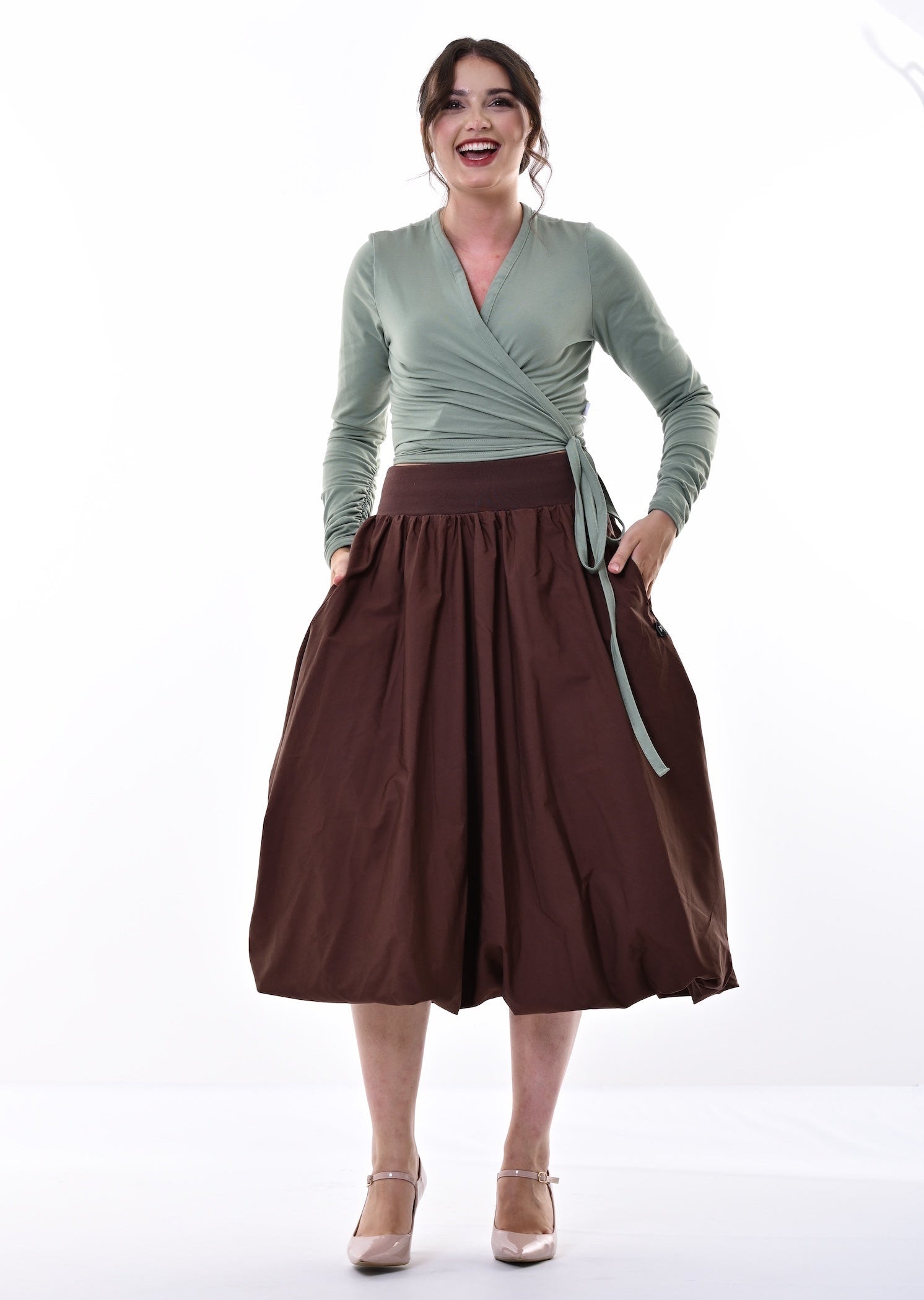 A woman modeling a sage green long-sleeve wrap top and a dark brown pleated midi skirt with side pockets, paired with beige heels, against a bright white studio background.