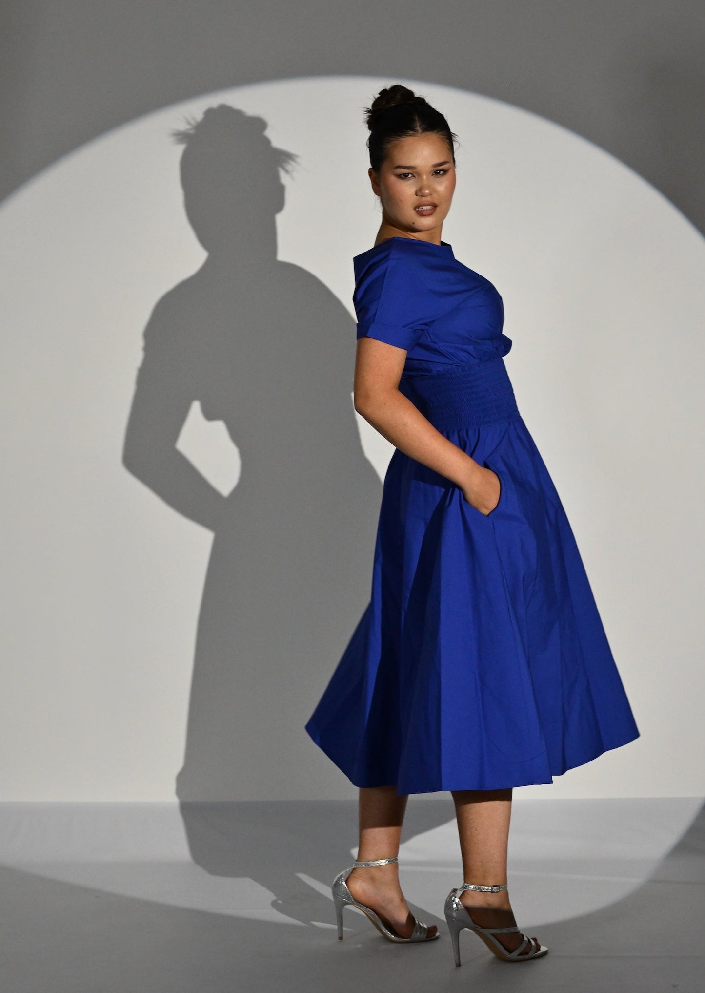 A woman models a royal blue midi dress with short sleeves, a draped neckline, and a flared skirt, standing in front of a spotlight on a gray studio floor that casts her shadow.
