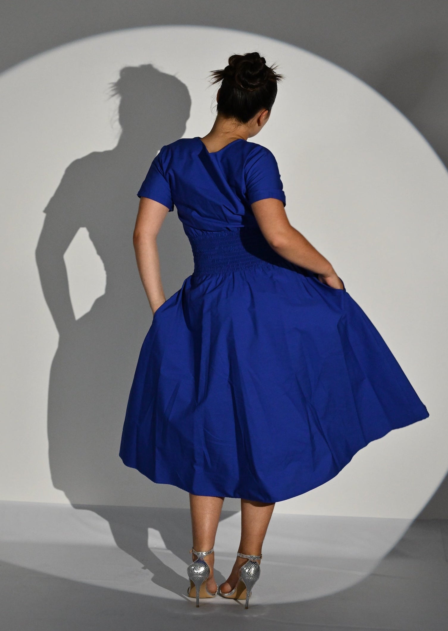 A woman wearing a bright royal blue short-sleeve midi dress is shown from behind, holding out the skirt to display its volume. She stands inside a large circular spotlight on a gray floor, casting a long shadow on the white spotlight surface.