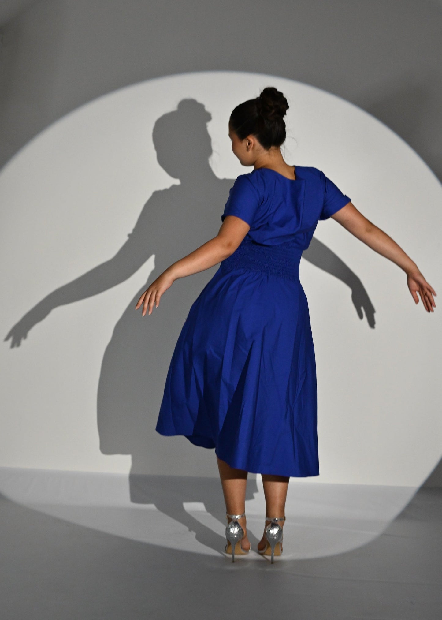 A woman stands with her back to the camera wearing a bright royal blue short-sleeve dress, posed inside a large circular spotlight on a white studio wall that casts a long shadow. She is wearing metallic silver high heels.