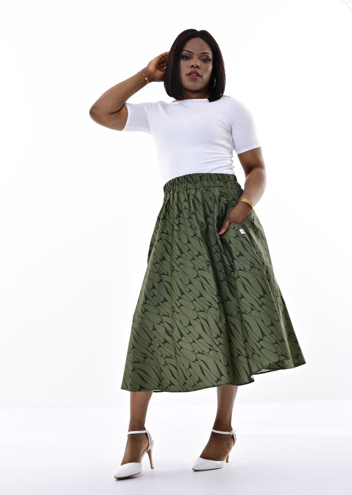 A woman modeling a high-waisted olive green midi skirt with a subtle abstract pattern, paired with a white t-shirt and white heels, against a bright white studio background.