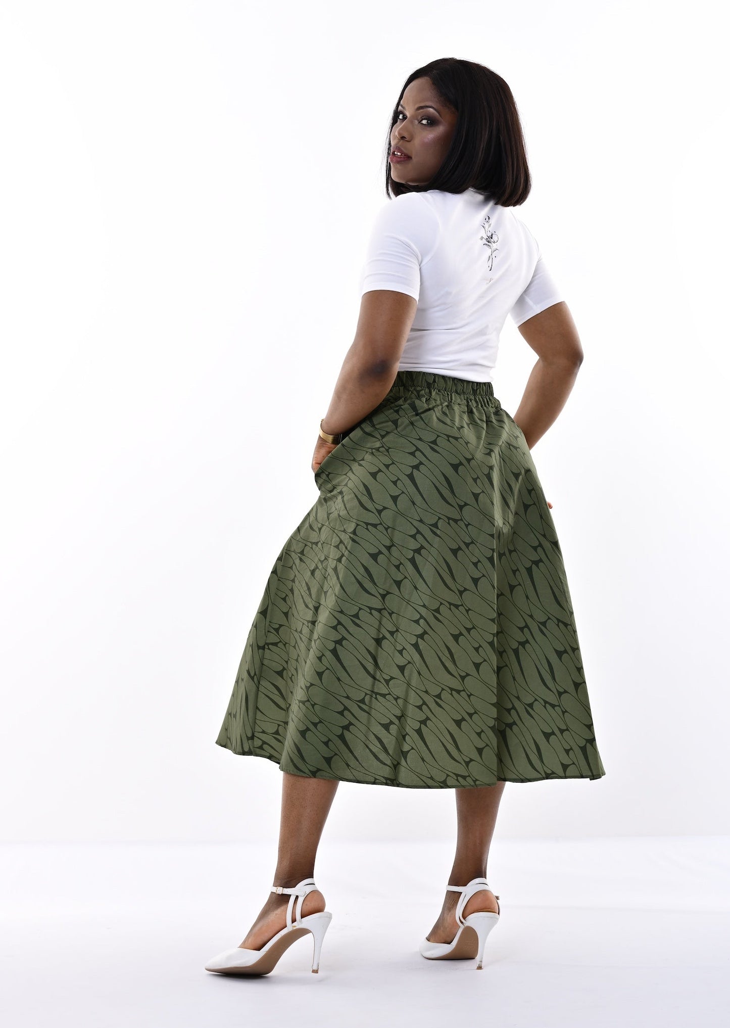 A woman stands in a studio wearing a white short-sleeve top and a high-waisted olive green midi skirt with a tonal abstract pattern. She is posed from the back, looking over her shoulder, wearing white high-heeled sandals against a bright white background.